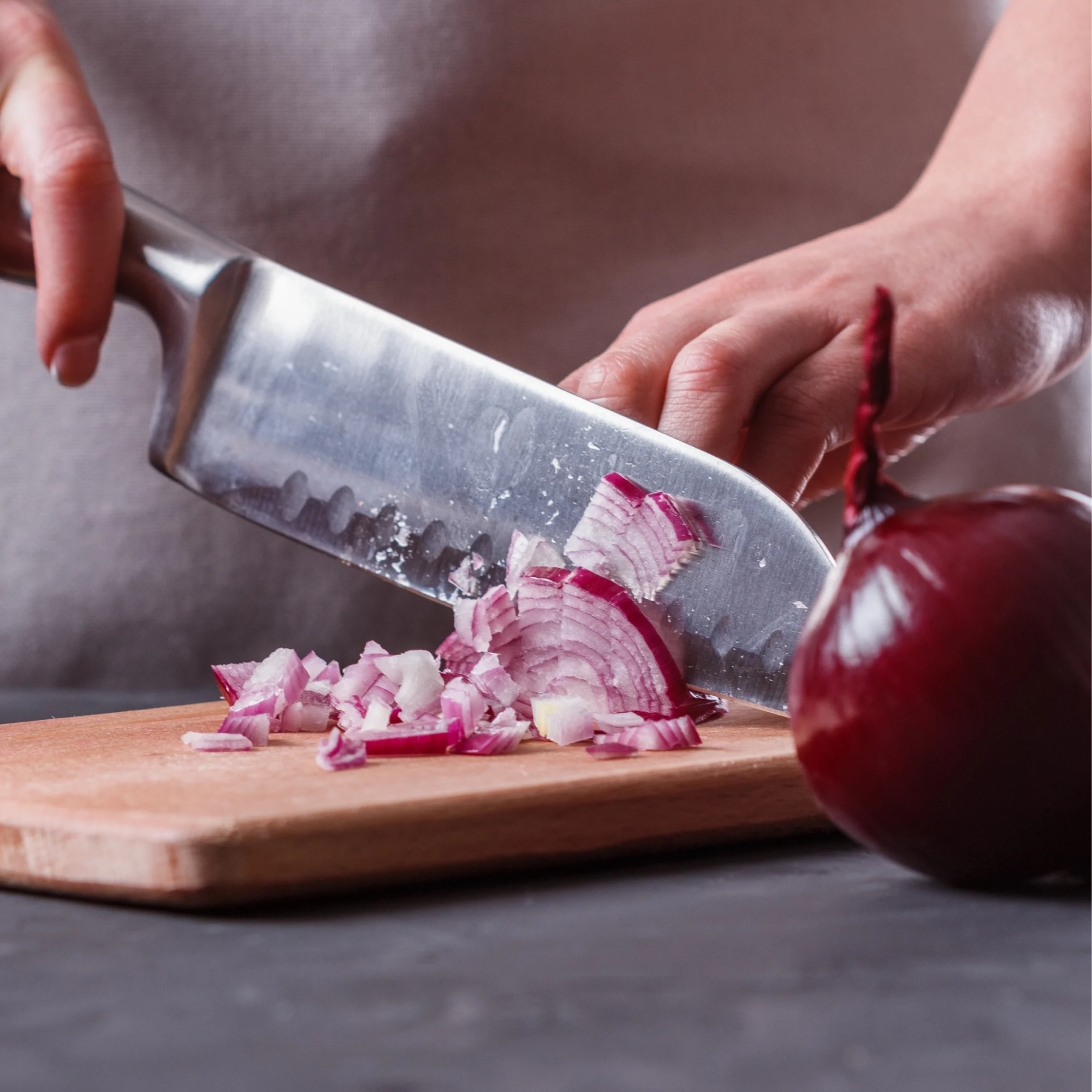 Close-up of a chef's hand slicing a red onion on a wooden cutting board with a stainless knife.