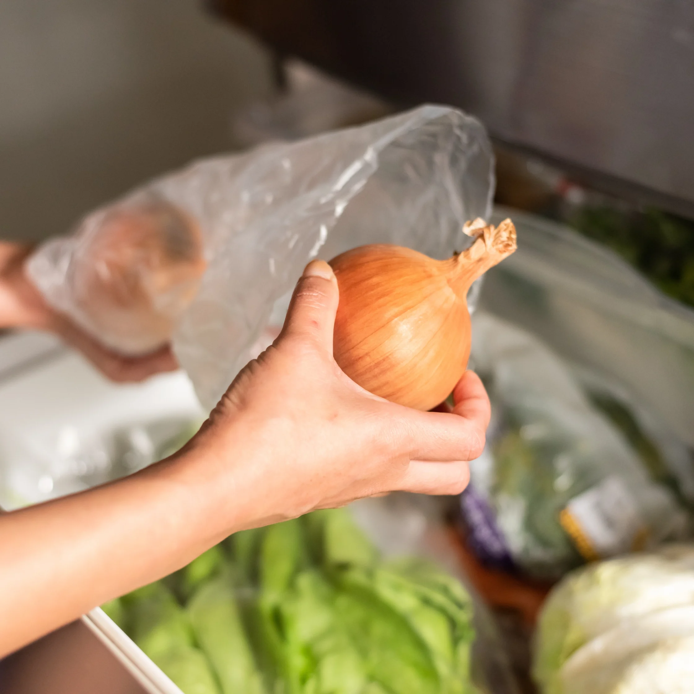 Person holding an onion with a peeled skin in a plastic bag in the background, kitchen scene blur behind.