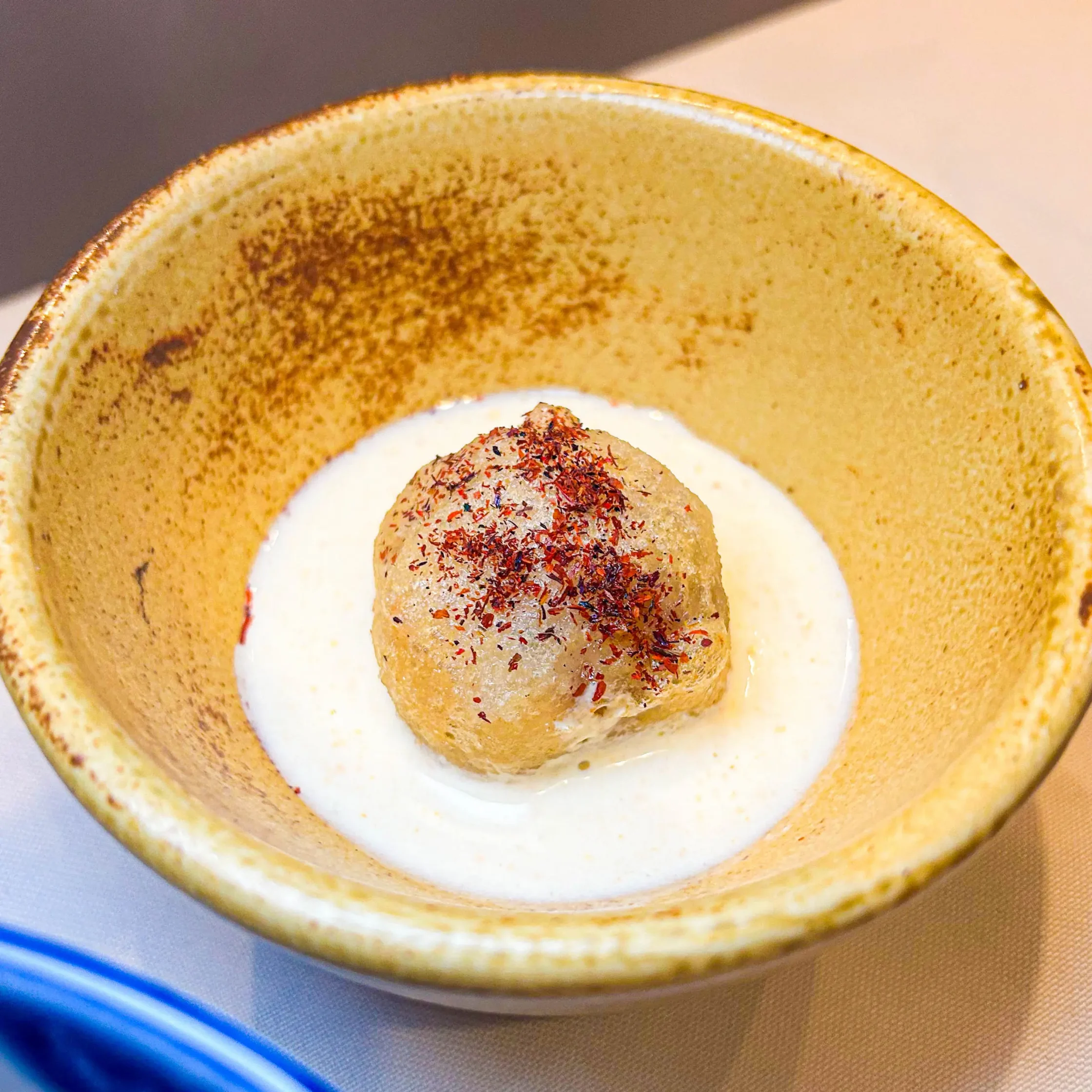 Close-up of a round, spice-coated ball resting on white sauce in a beige ceramic bowl, with a blue utensil at the bottom edge.