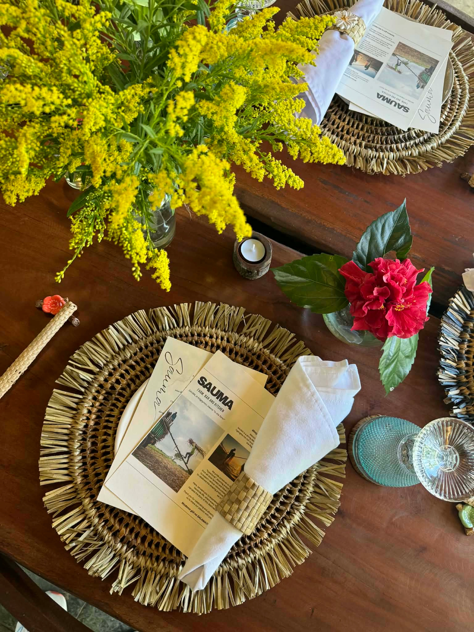 Elegant tropical table setting with woven placemats, yellow flowers, and a red hibiscus centerpiece; menu and napkin with napkin ring visible.