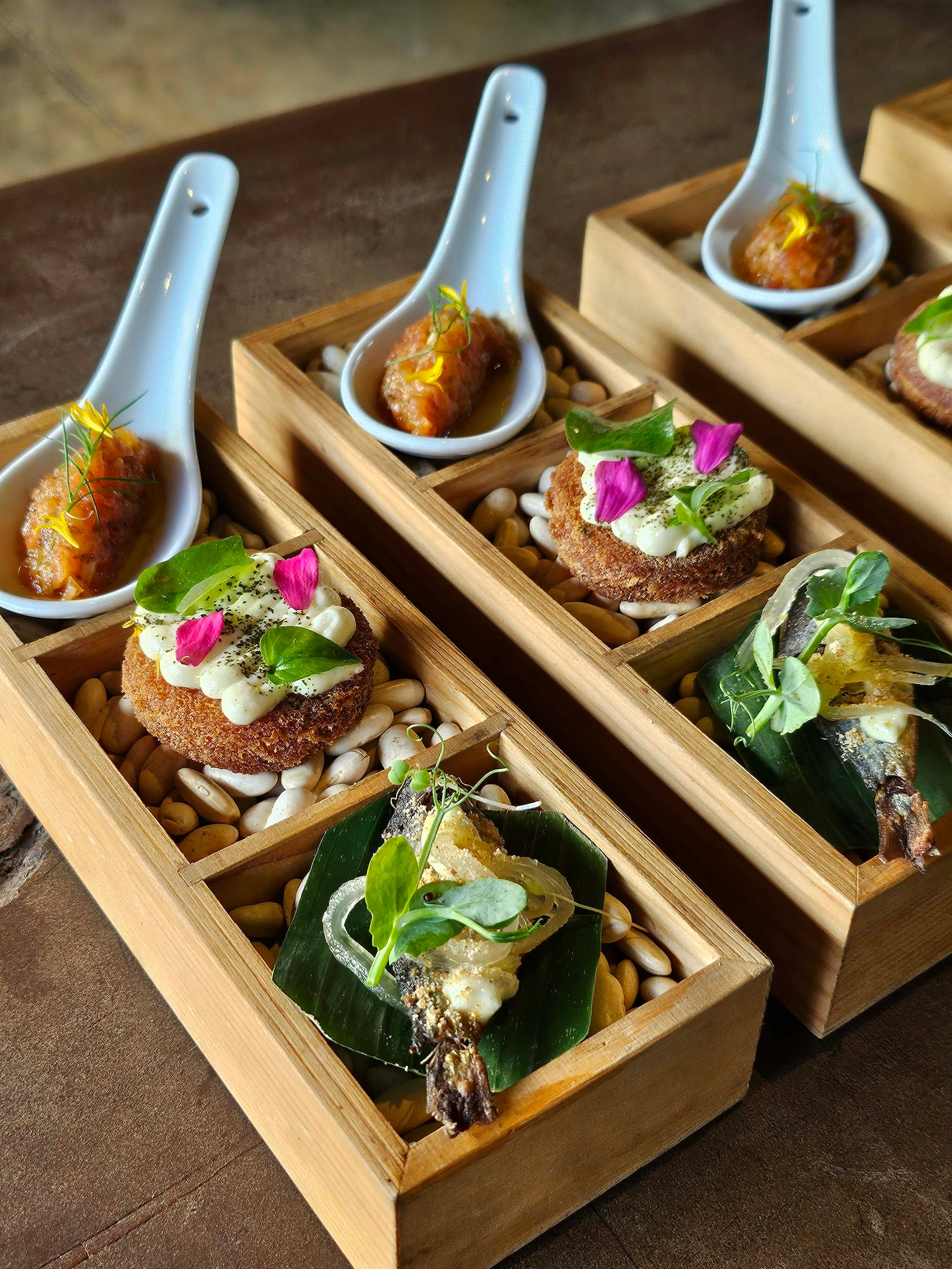 Assorted tapas in wooden boxes with cobblestone bed: croquettes topped with cream, greens, and pink petals; small spoons with fried bites in each compartment.