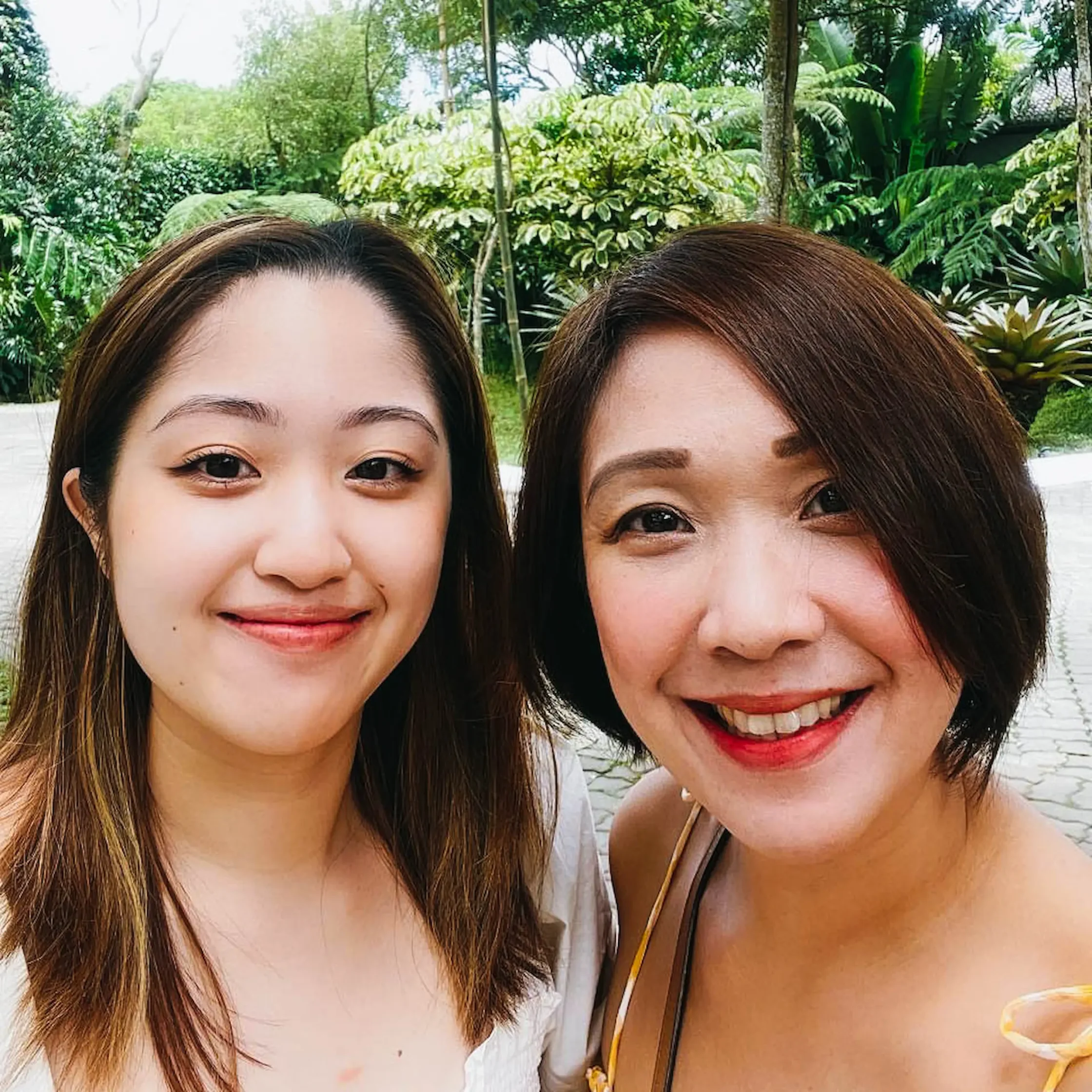 Two women smiling in a sunny garden with tropical plants in the background.