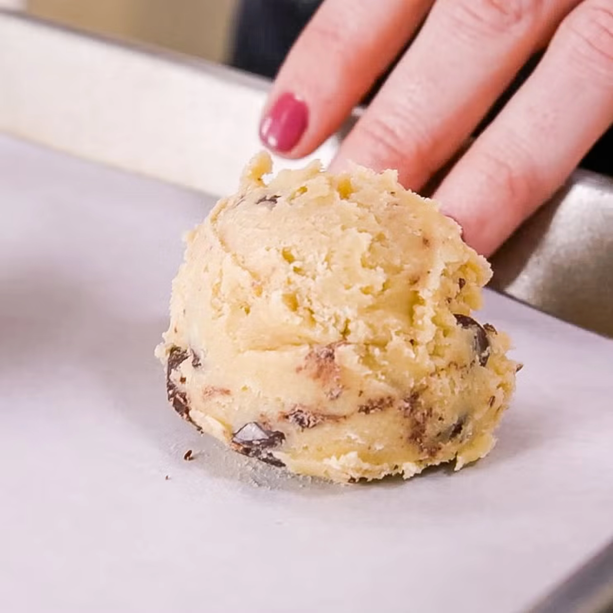 Close-up of a hand with pink nail polish pinching a crumbly ball of cookie dough.