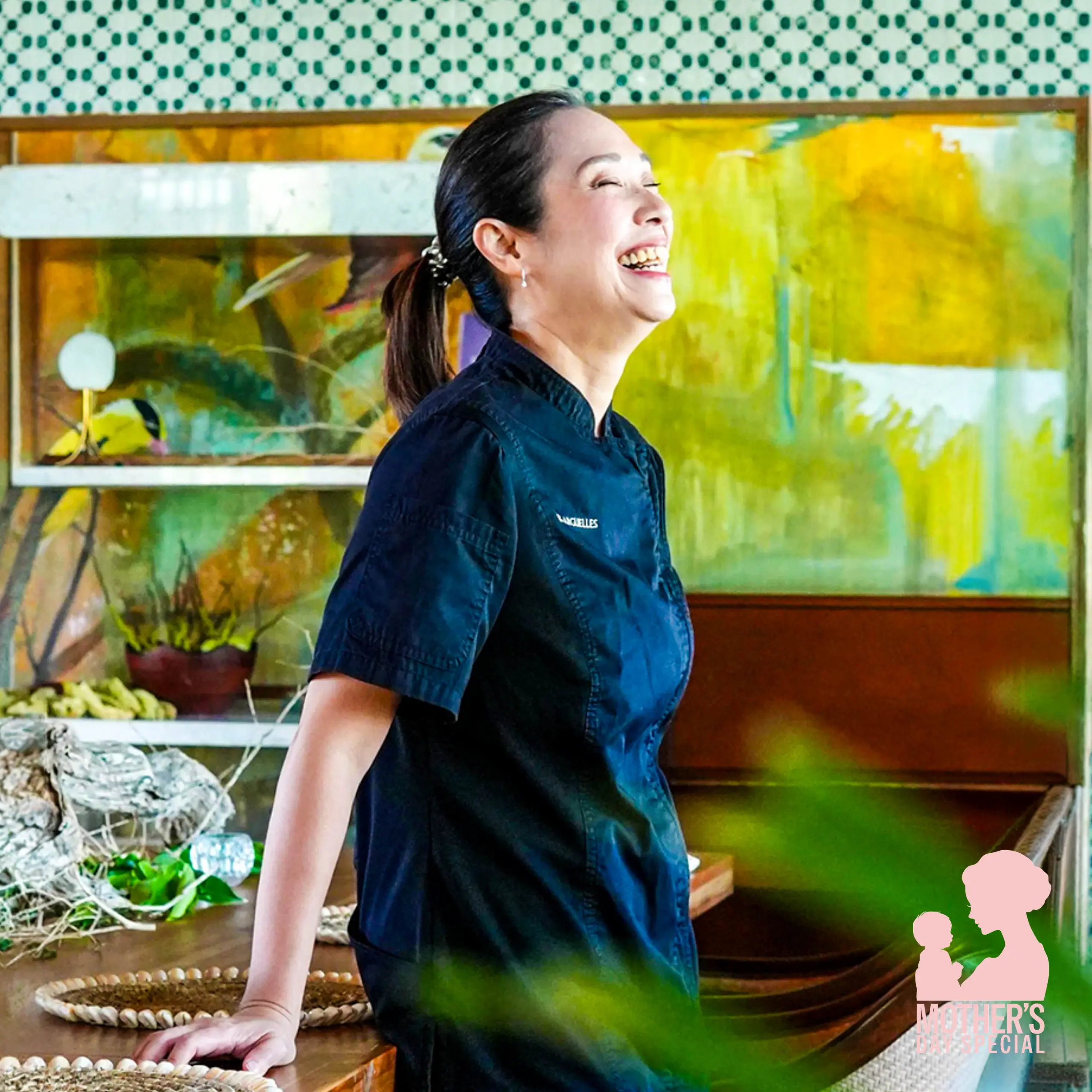 Woman in a dark chef jacket smiles while standing beside a colorful mural and produce display in a kitchen-like setting; pink 'Mother's Day Special' graphic on bottom right.