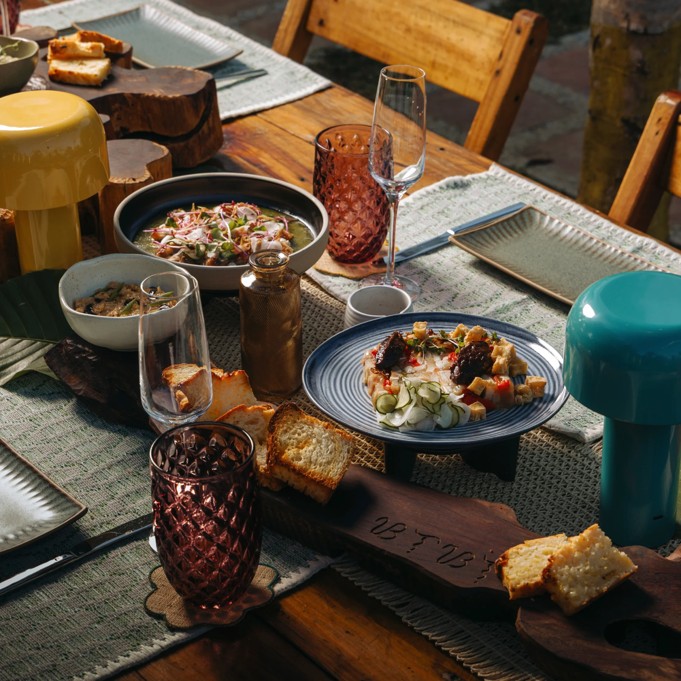 A rustic dining table set with plates of food, bread, red glassware, and a clear wine glass on a textured placemat outside.