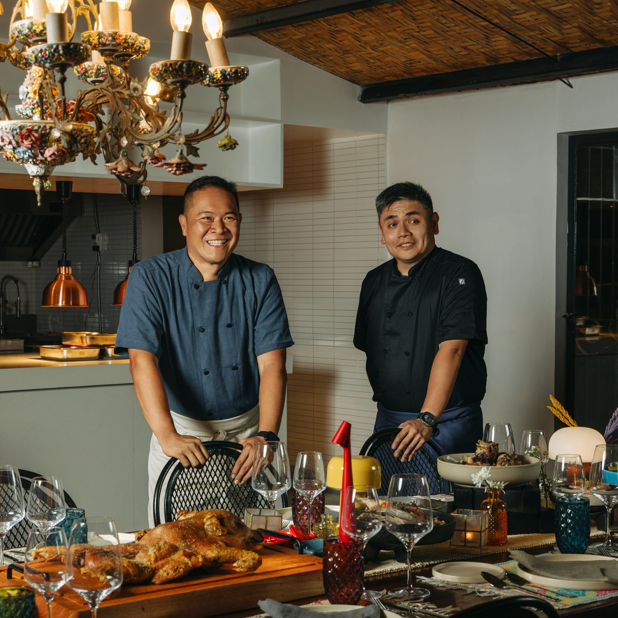 Two male chefs stand at a kitchen island, smiling, with a table of bread, drinks, and place settings in front of them. A decorative chandelier hangs above.