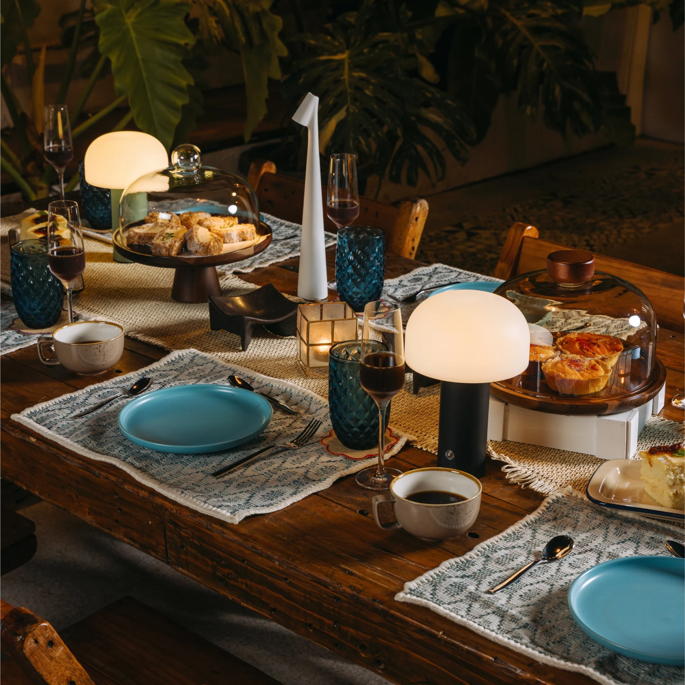 Table set for a dinner with blue dishes, glassware, and a cake under a glass cloche in warm lighting.
