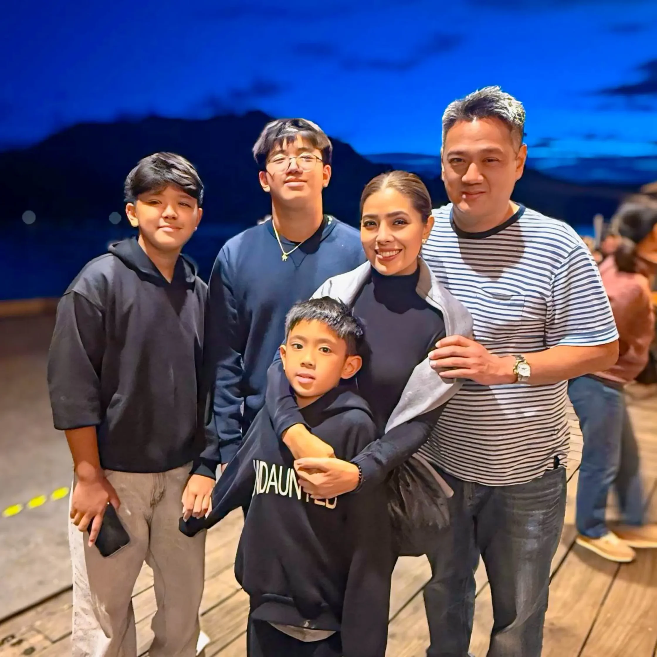 Group of four people posing for a photo on a wooden deck at night, dressed in casual dark clothing, smiling at the camera.
