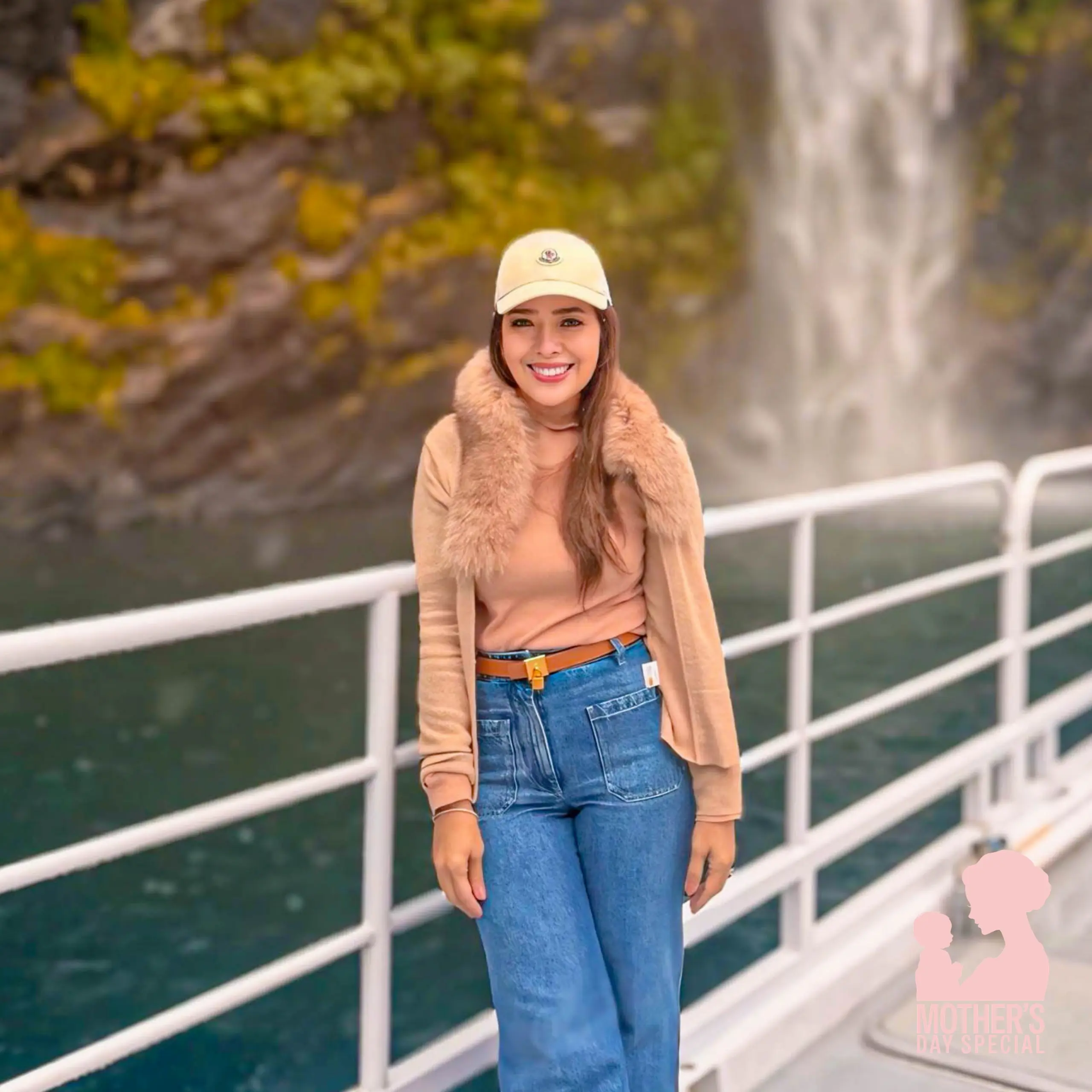 Smiling woman in a tan coat and beige cap standing on a railing with a waterfall in the background.