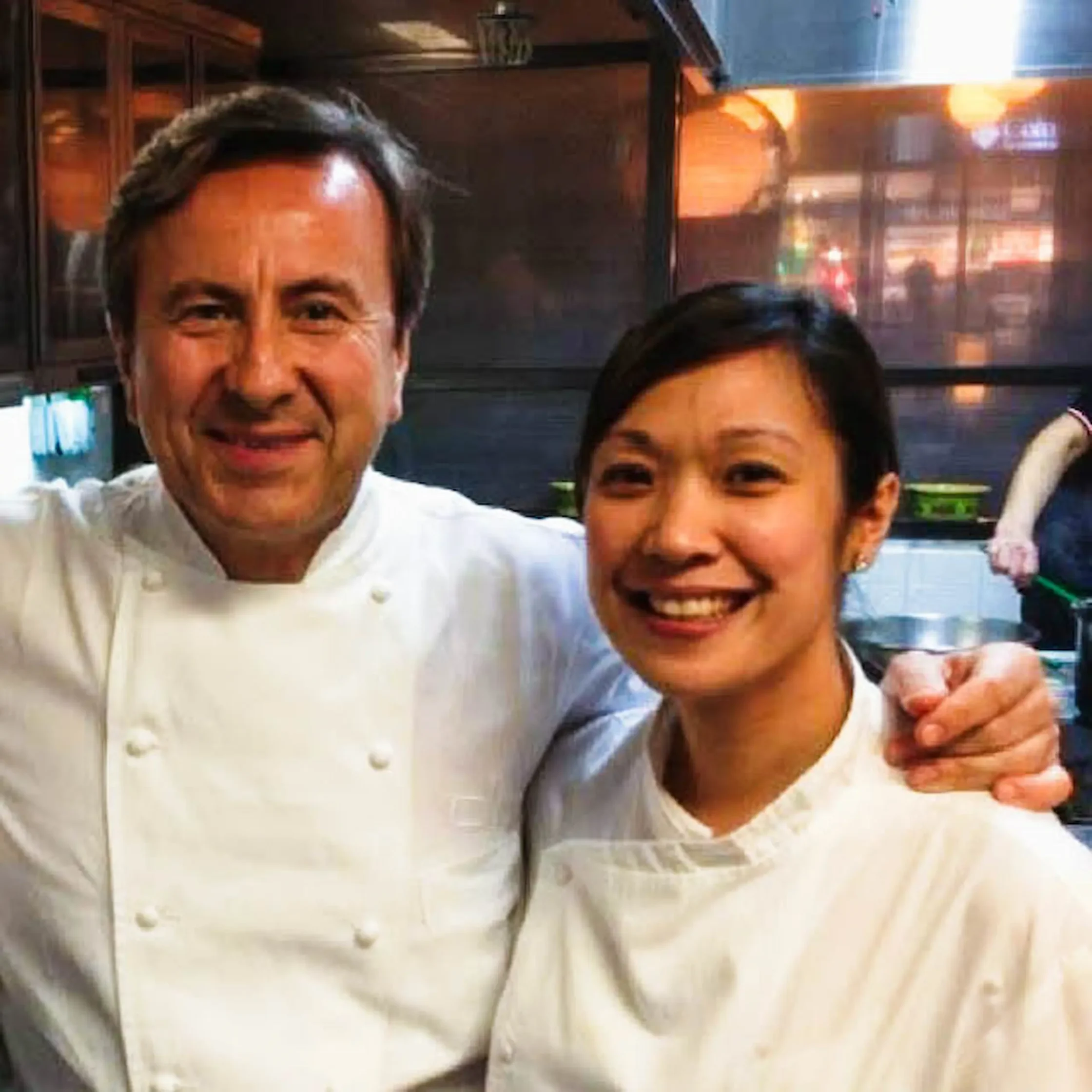 Two people smiling in white chef jackets, posing together in a restaurant or kitchen setting.