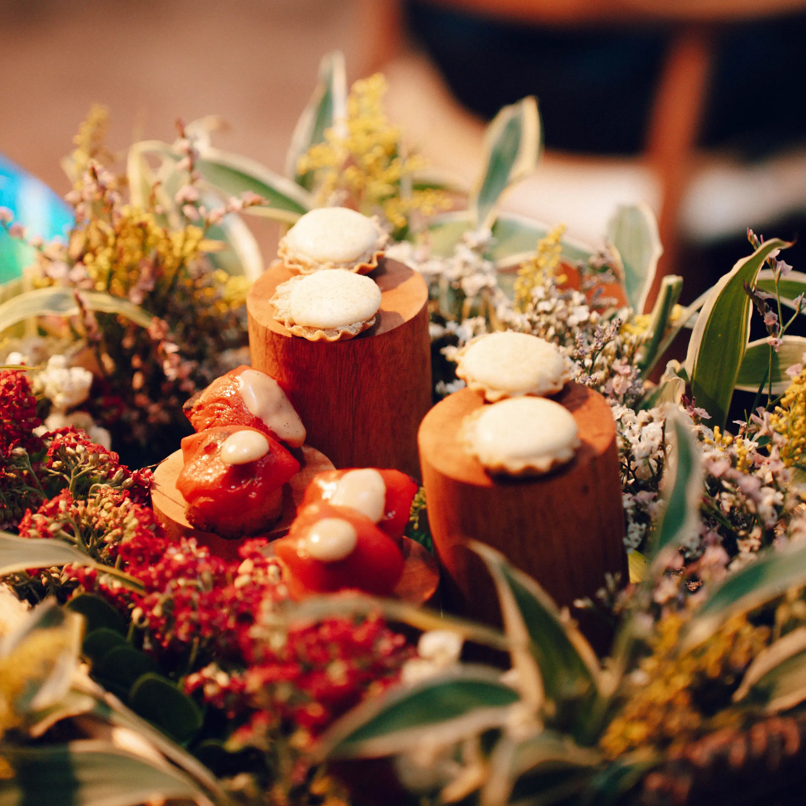 Small cream-topped pastries mounted on wooden bases among a vivid bed of dried flowers.