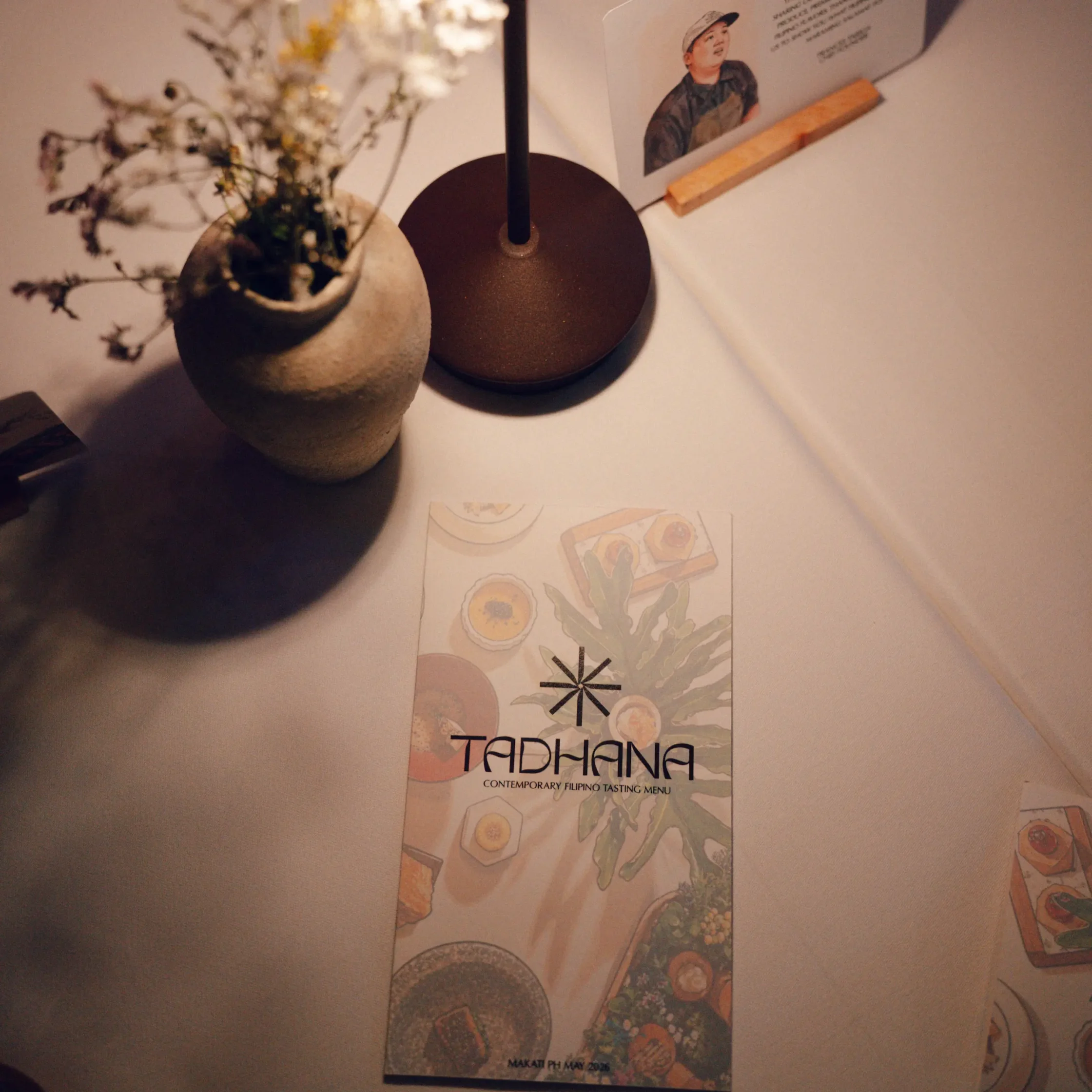 Tall vertical menu card on a white table showing botanical illustrations and the text 'Contemporary Filipino Tasting Menu'; a dark lamp base sits nearby with a potted plant in the background.