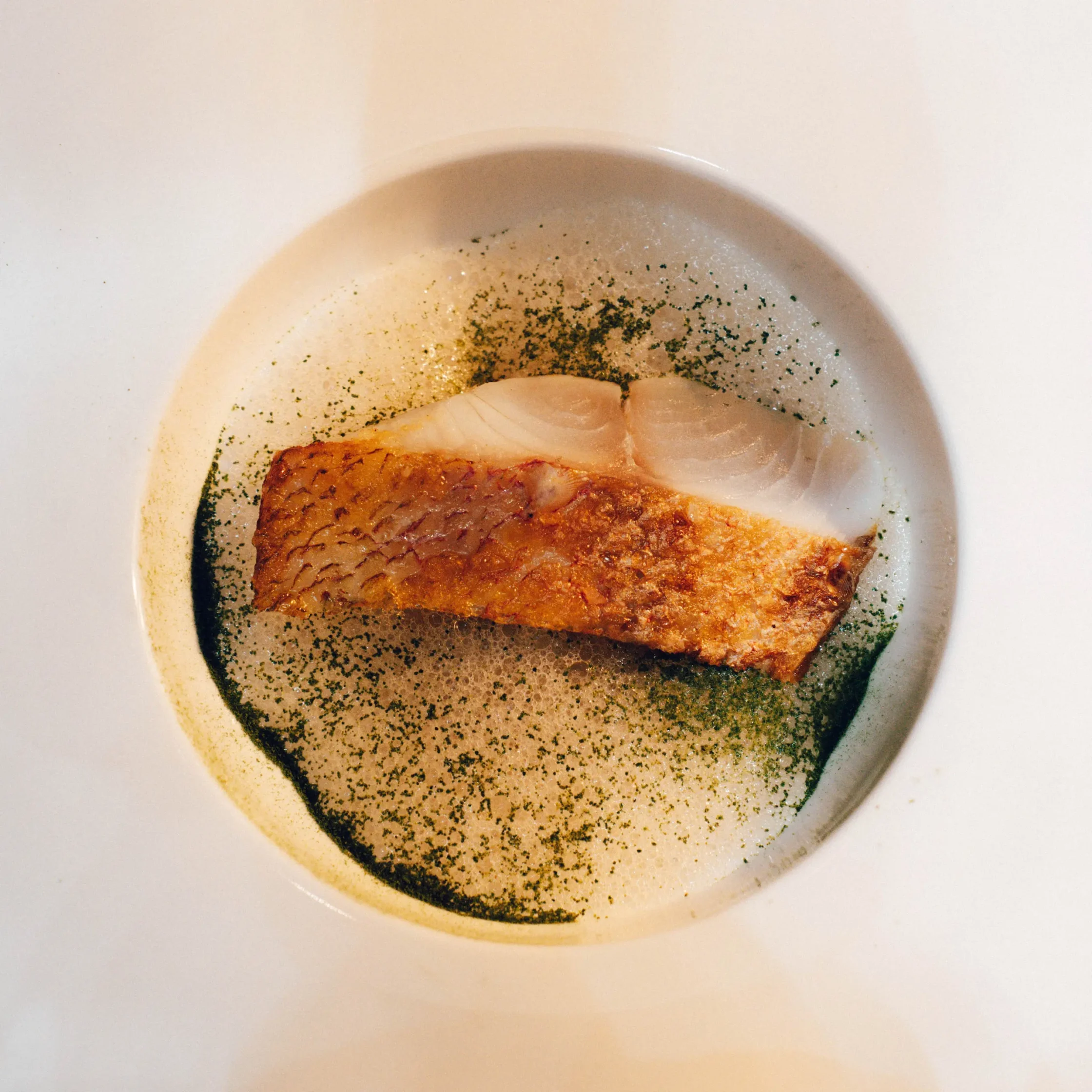Pan-seared fish fillet on a white plate with scattered green herb seasoning.
