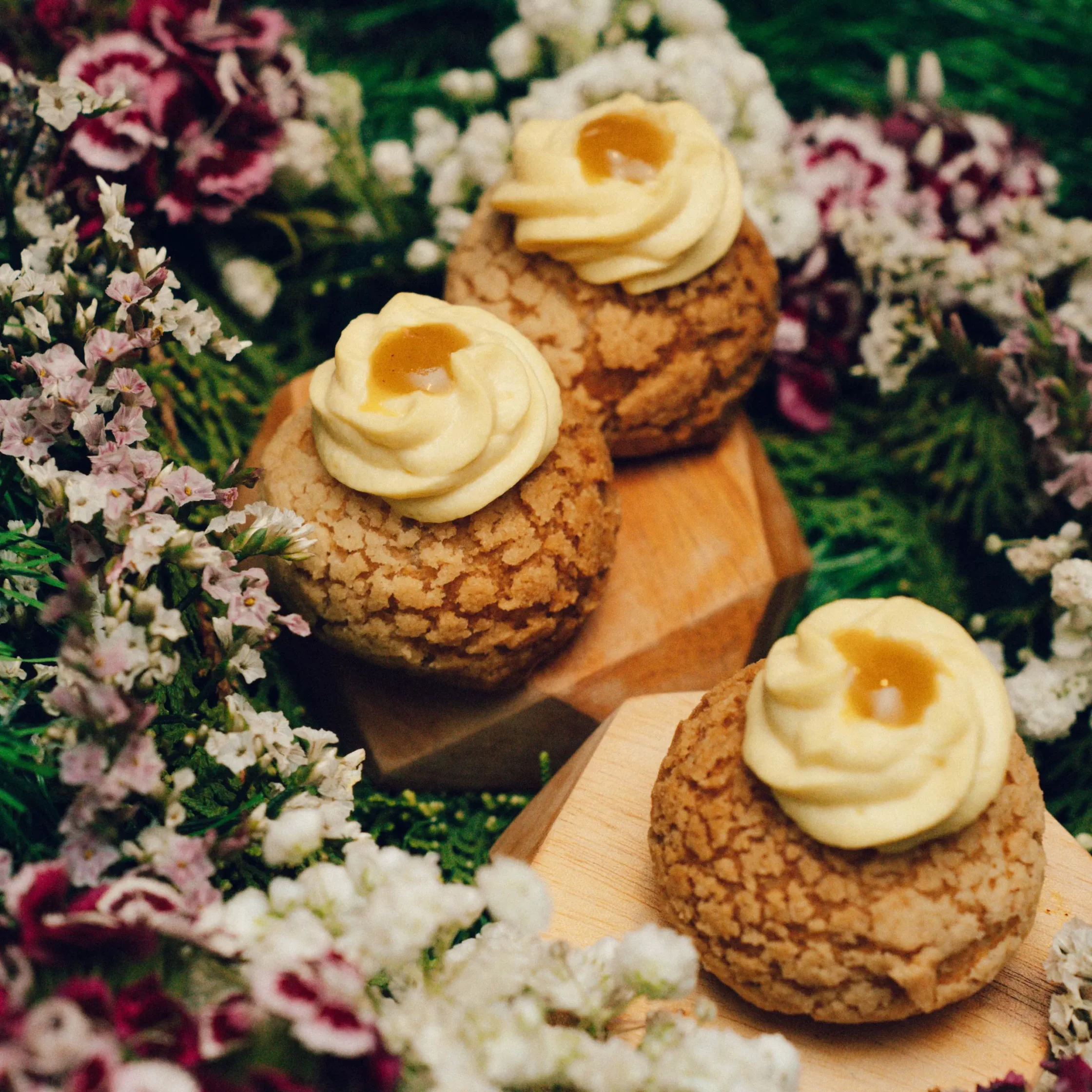 Crumbly round cookies topped with swirled vanilla frosting and a knob of jam, arranged on a wooden board among pink and white flowers.