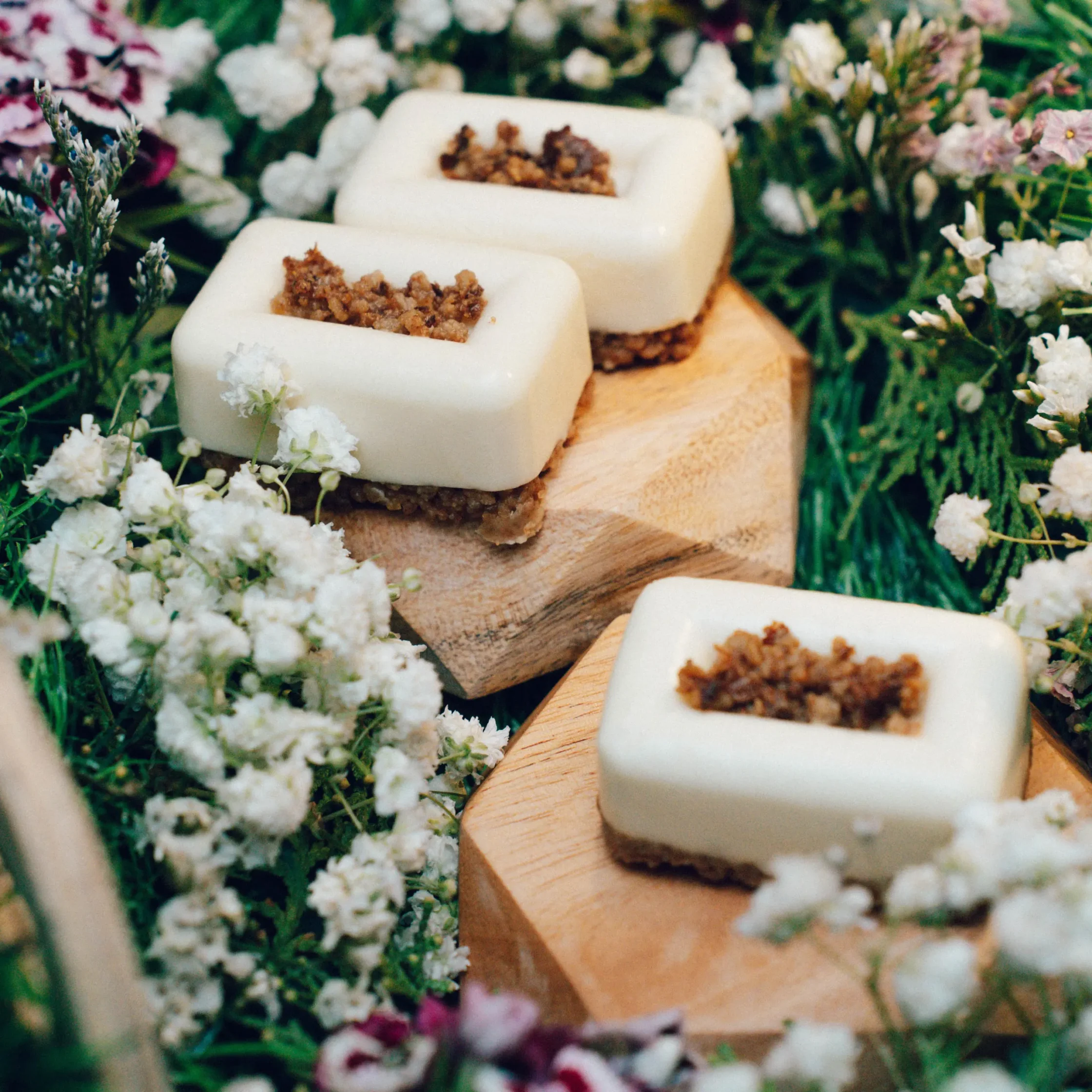 White rectangular soaps with brown sugar scrub, arranged on a wooden surface among white and pastel flowers.