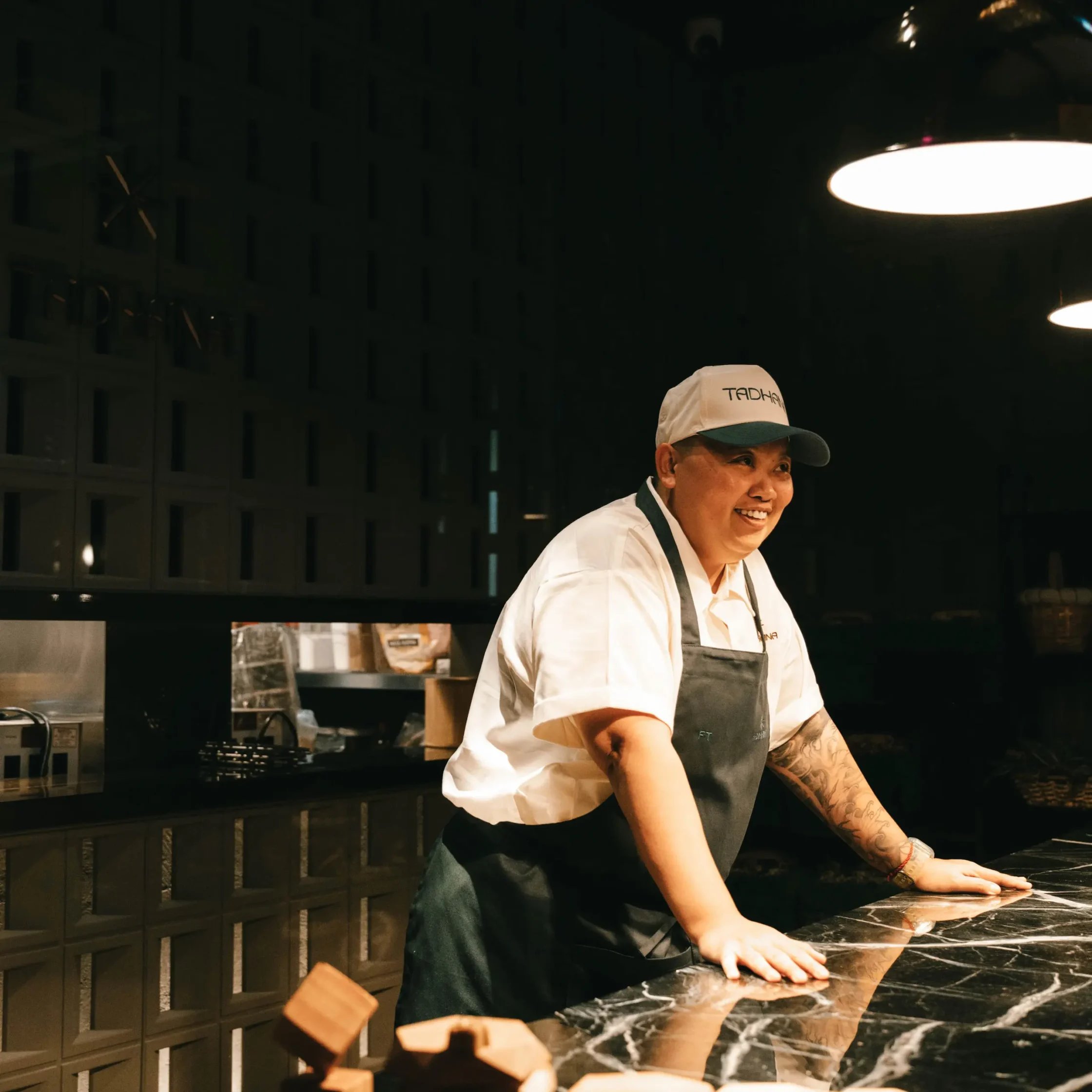 Chef in a white uniform and cap leaning on a marble kitchen counter, smiling in a dimly lit kitchen.