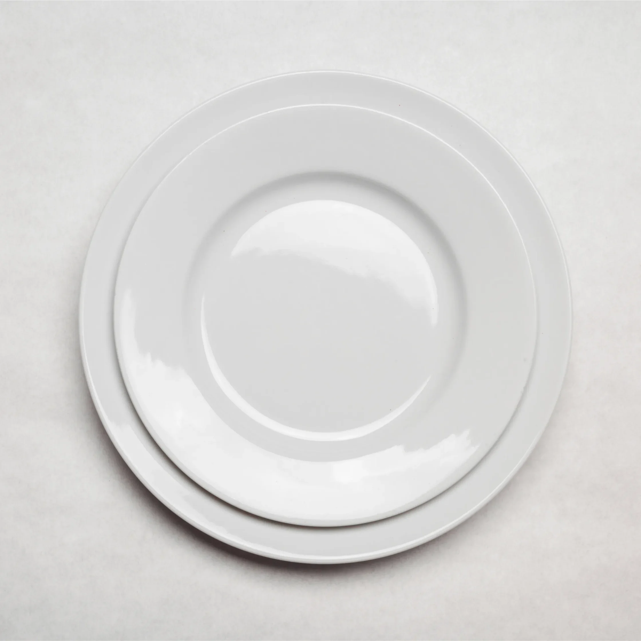 Small white bread and butter plate stacked on a larger white plate, photographed against a clean white background, commonly used for serving bread rolls, butter, and small accompaniments.