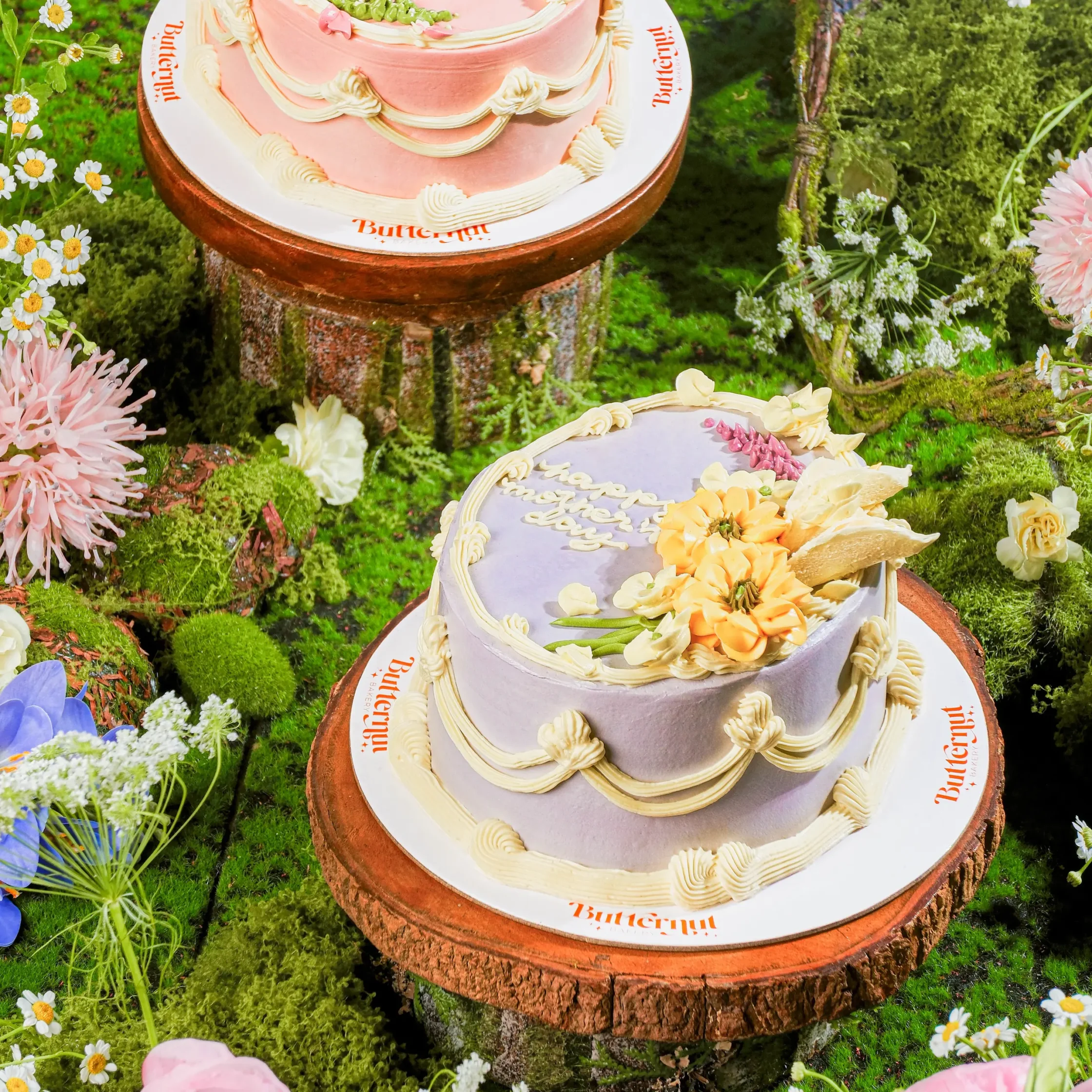 Outdoor display of pastel pink and lavender cakes on wooden tree-stump stands amid moss and flowers.