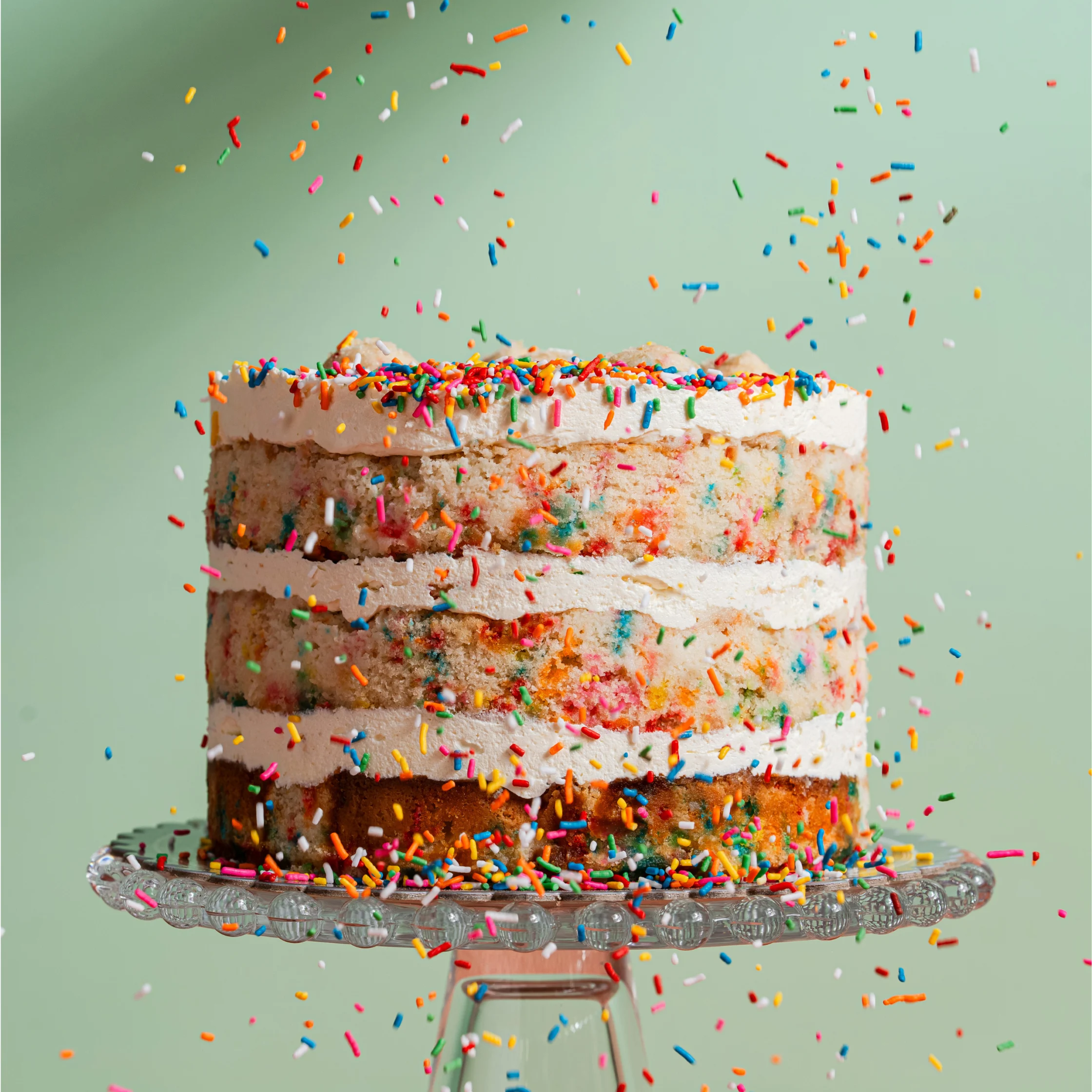 Three-layer confetti cake with vanilla frosting and rainbow sprinkles falling around the cake on a glass stand.