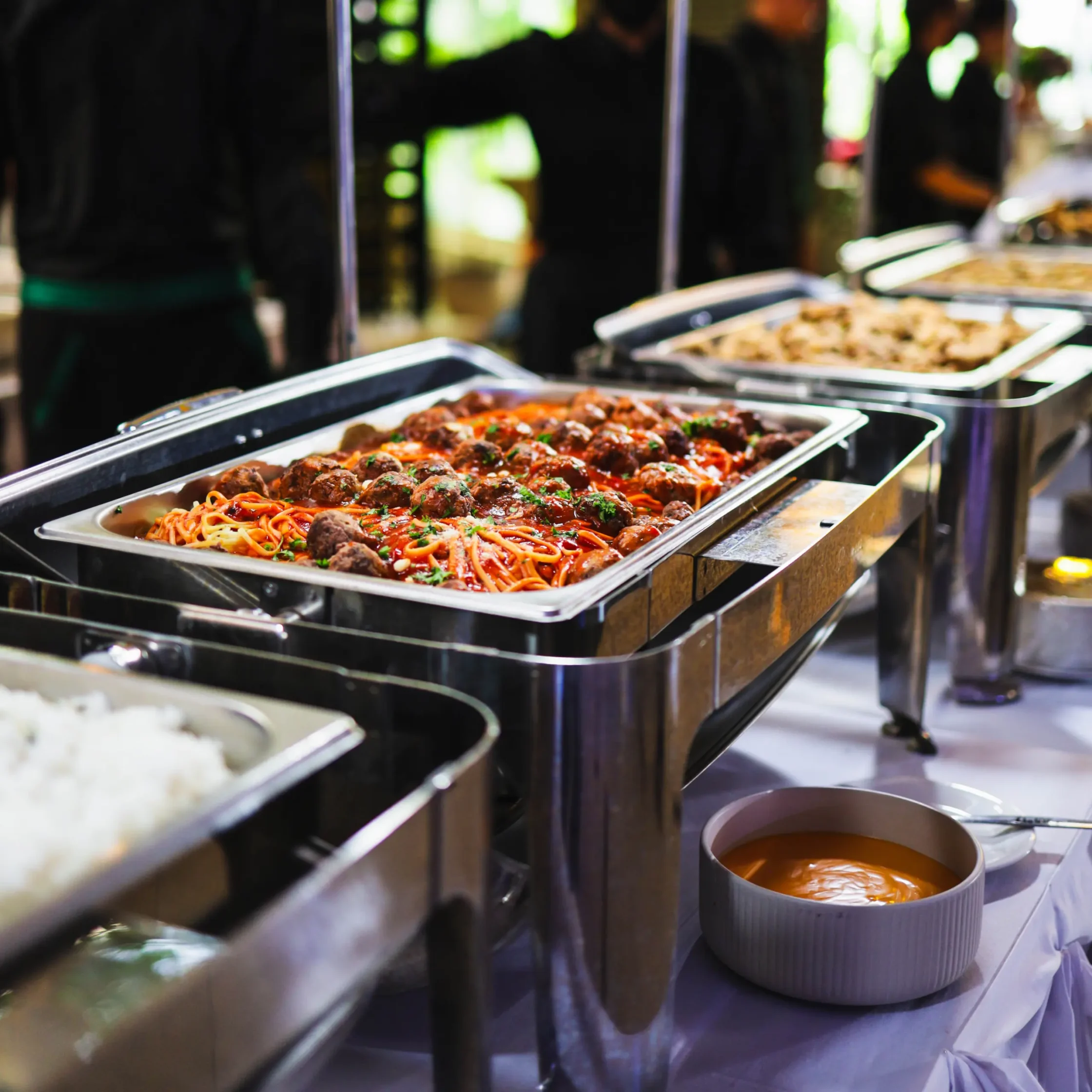 Buffet serving station with trays of pasta and meatballs in the foreground and chefs blurred in the background.