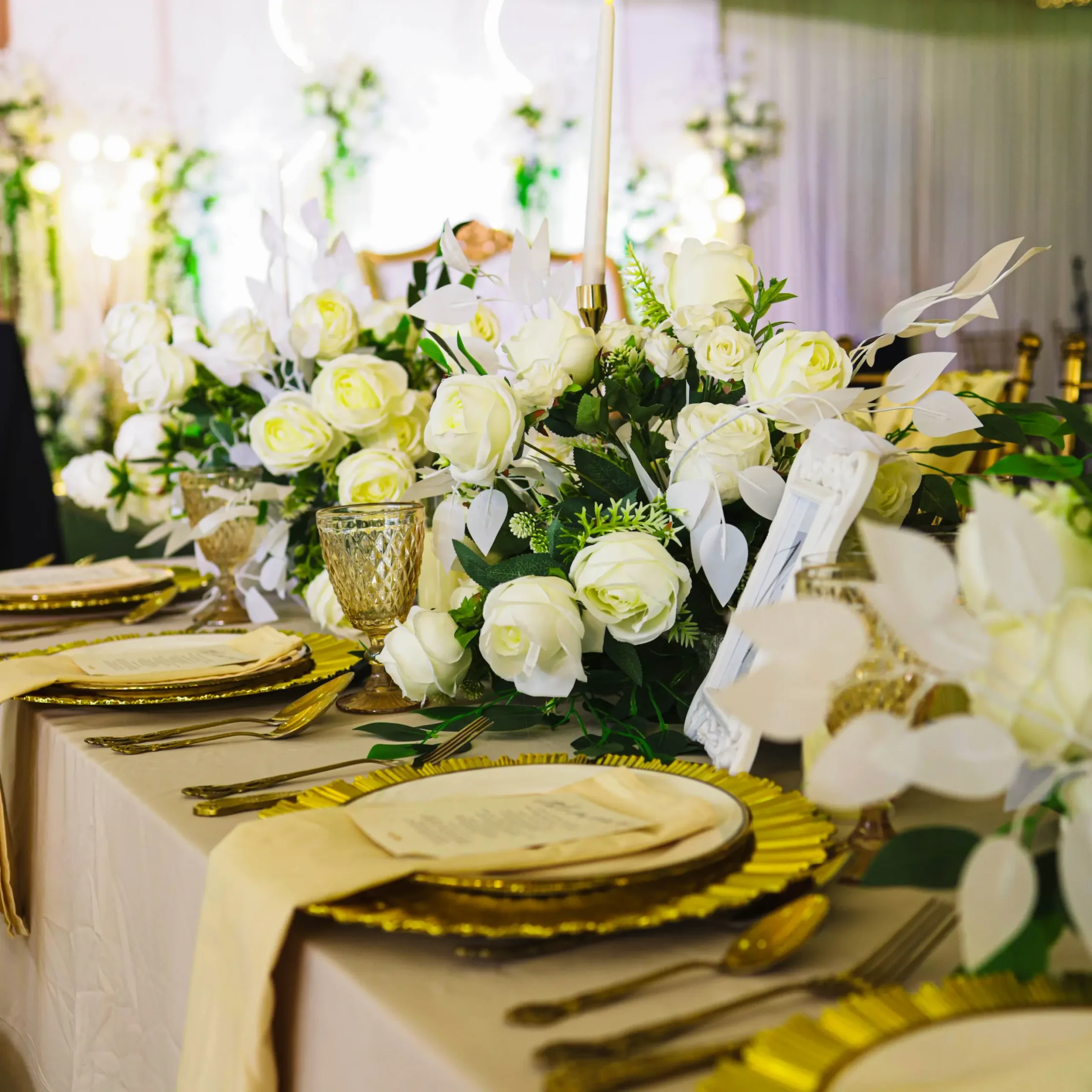 Elegant banquet table with white roses, greenery, and gold utensils centerpiece. Enjoying a formal event setup.