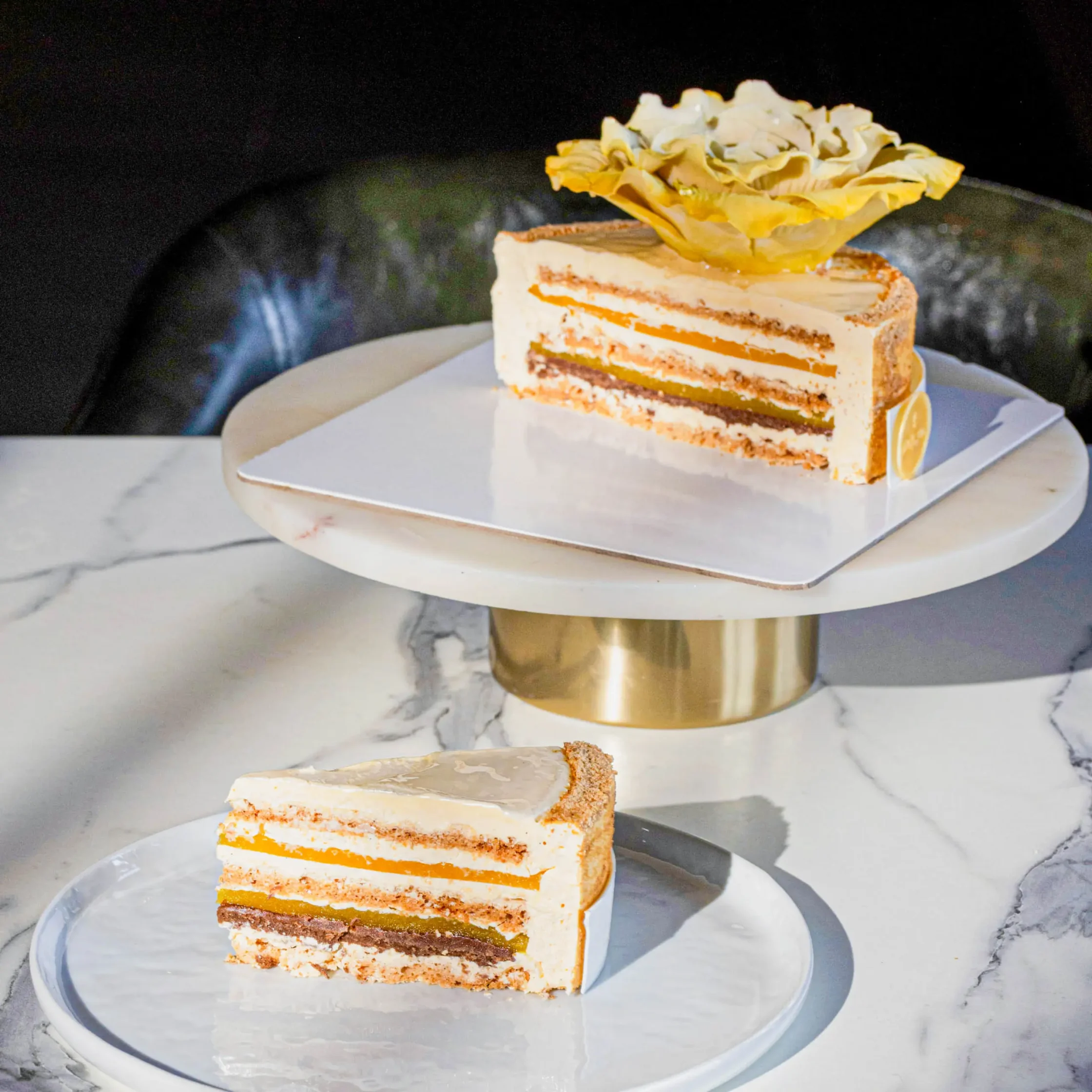 Layered vanilla cake slice with orange jam filling and cream frosting, topped with a yellow flower on a white plate.