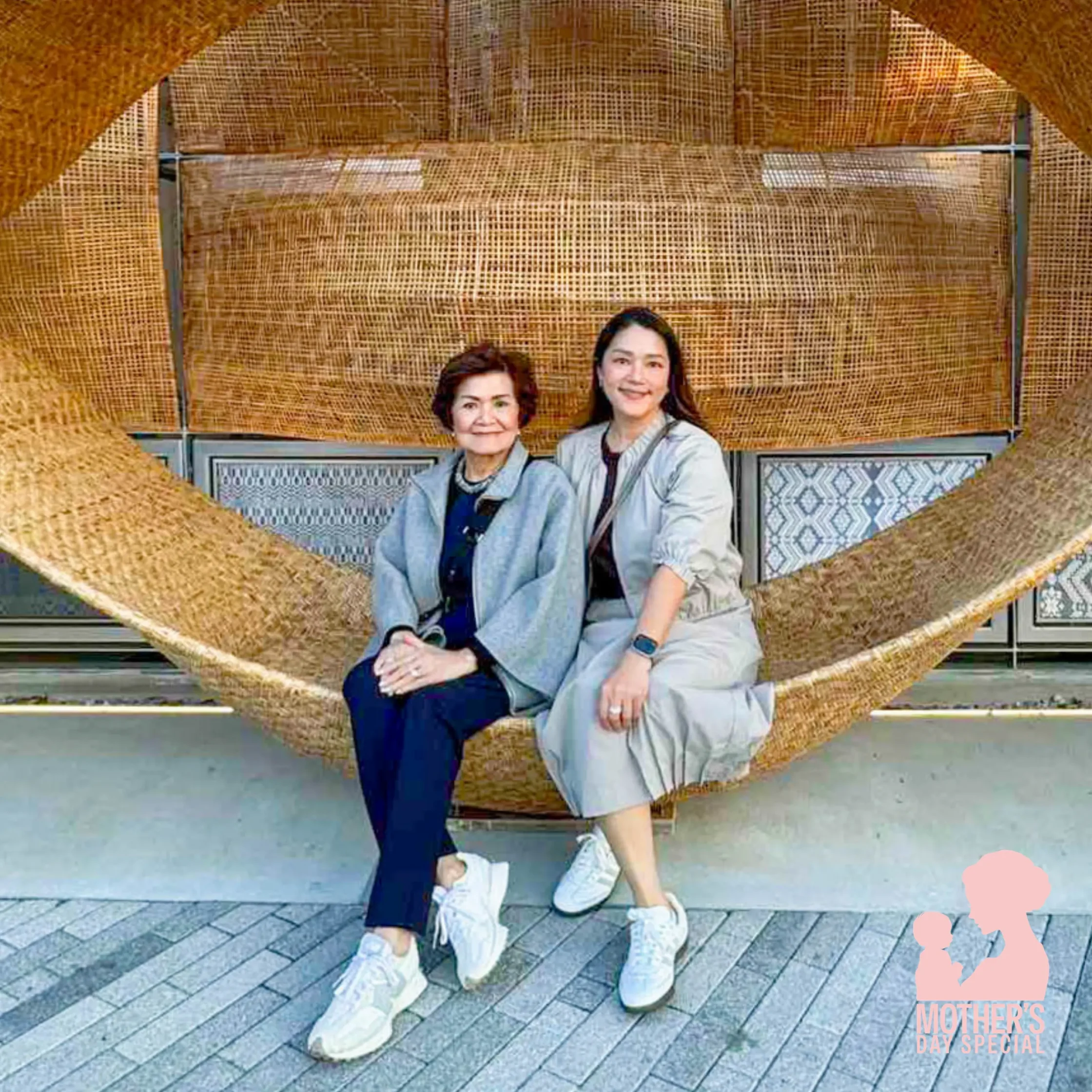 Two women sit closely on a giant woven wicker sculpture, smiling at the camera outdoors.
