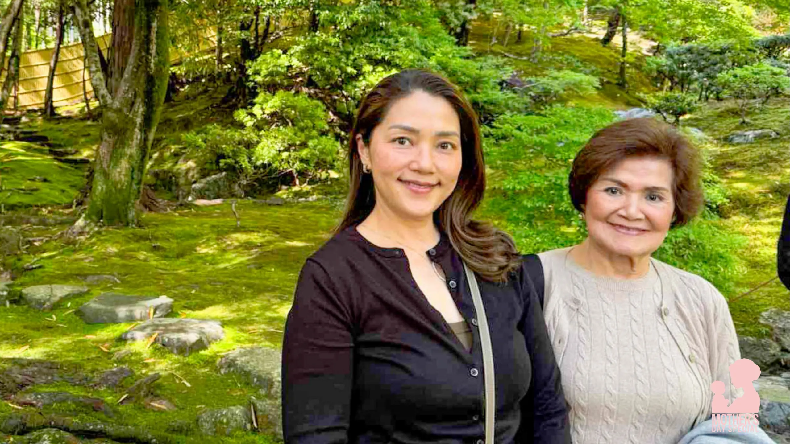 Two women posing and smiling in a lush green garden with mossy ground and trees behind them.