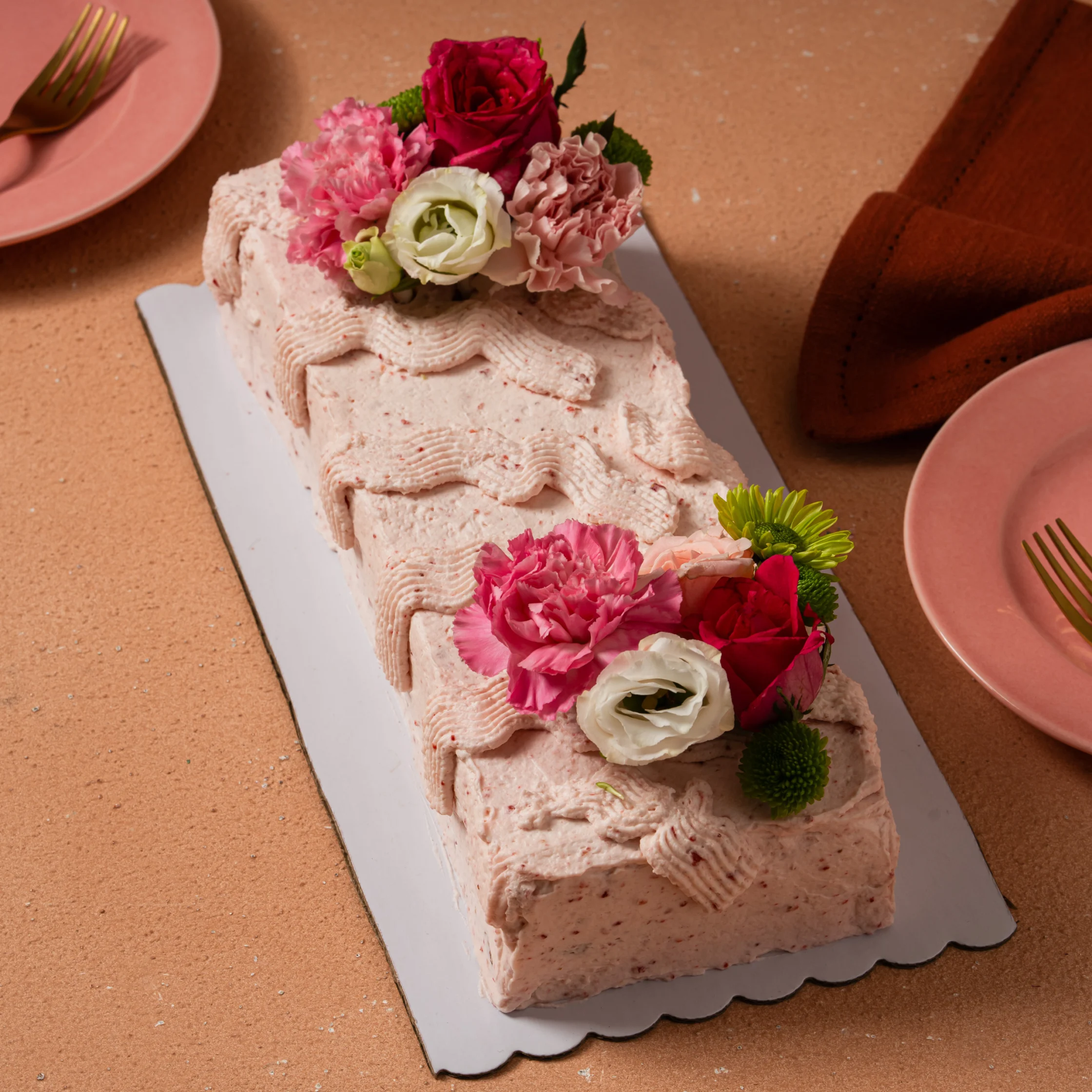 Rectangular strawberry shortcake with pink frosting and floral decorations on a cake board beside pink plates.