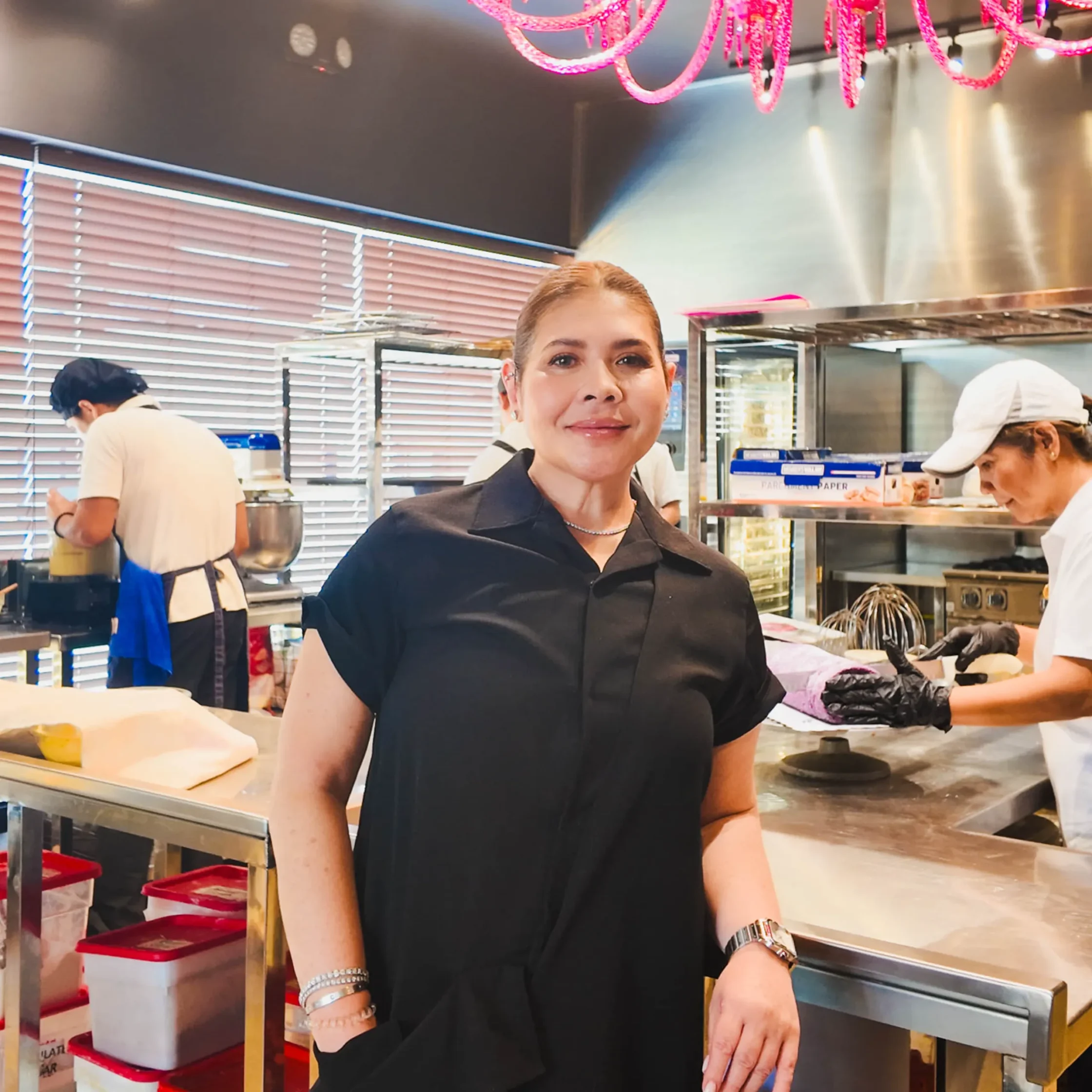 Trish Lacson posing inside 50&Co Bakery production kitchen while bakery staff decorate cakes and prepare desserts, showcasing professional baking operations and artisan pastry craftsmanship.