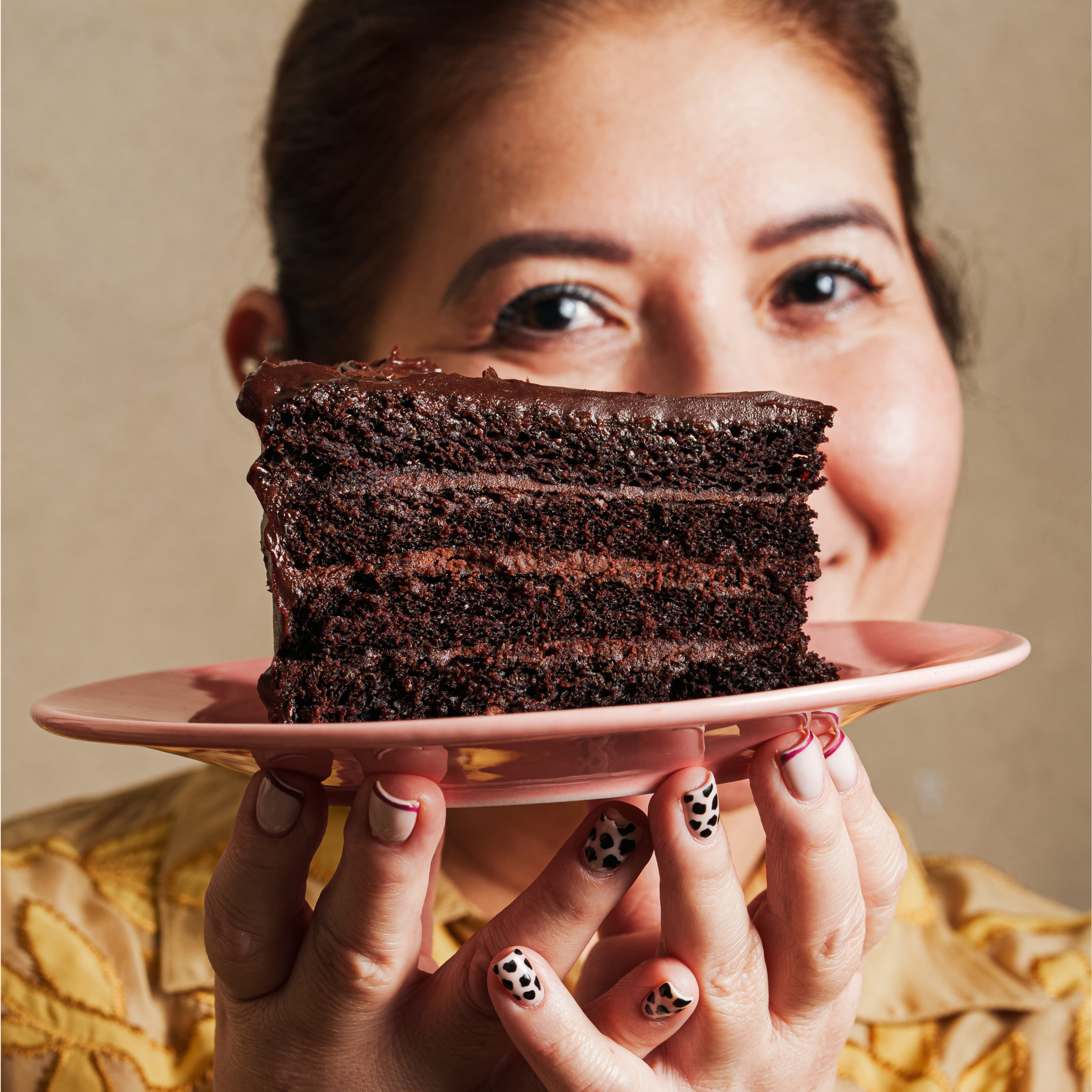Woman holding a slice of rich chocolate layer cake on a pink plate close to the camera.