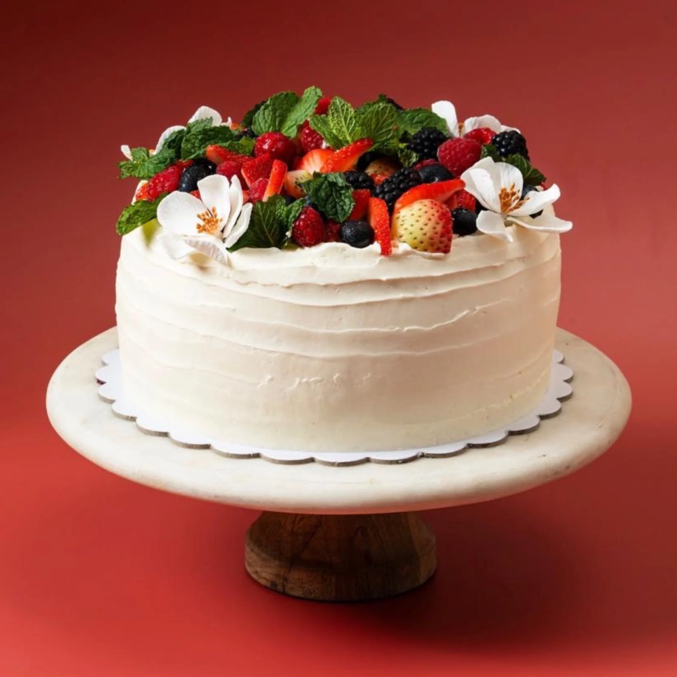 White Vanilla Chantilly cake decorated with strawberries, raspberries, blueberries, mint leaves, and white flowers on a cake stand.