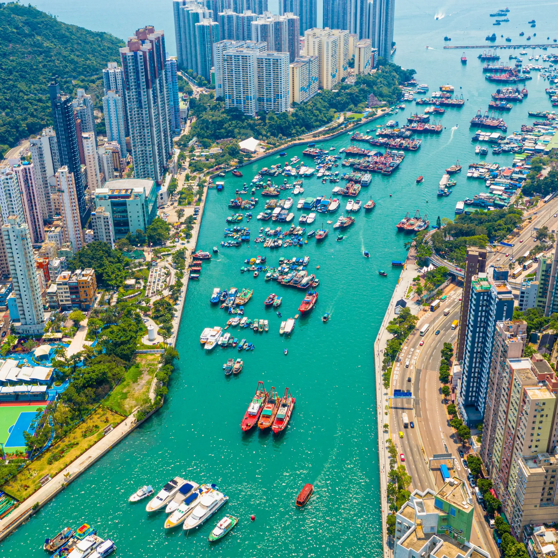 Aerial view of a coastal city with densely packed high-rise buildings along a turquoise harbor filled with boats and yachts.