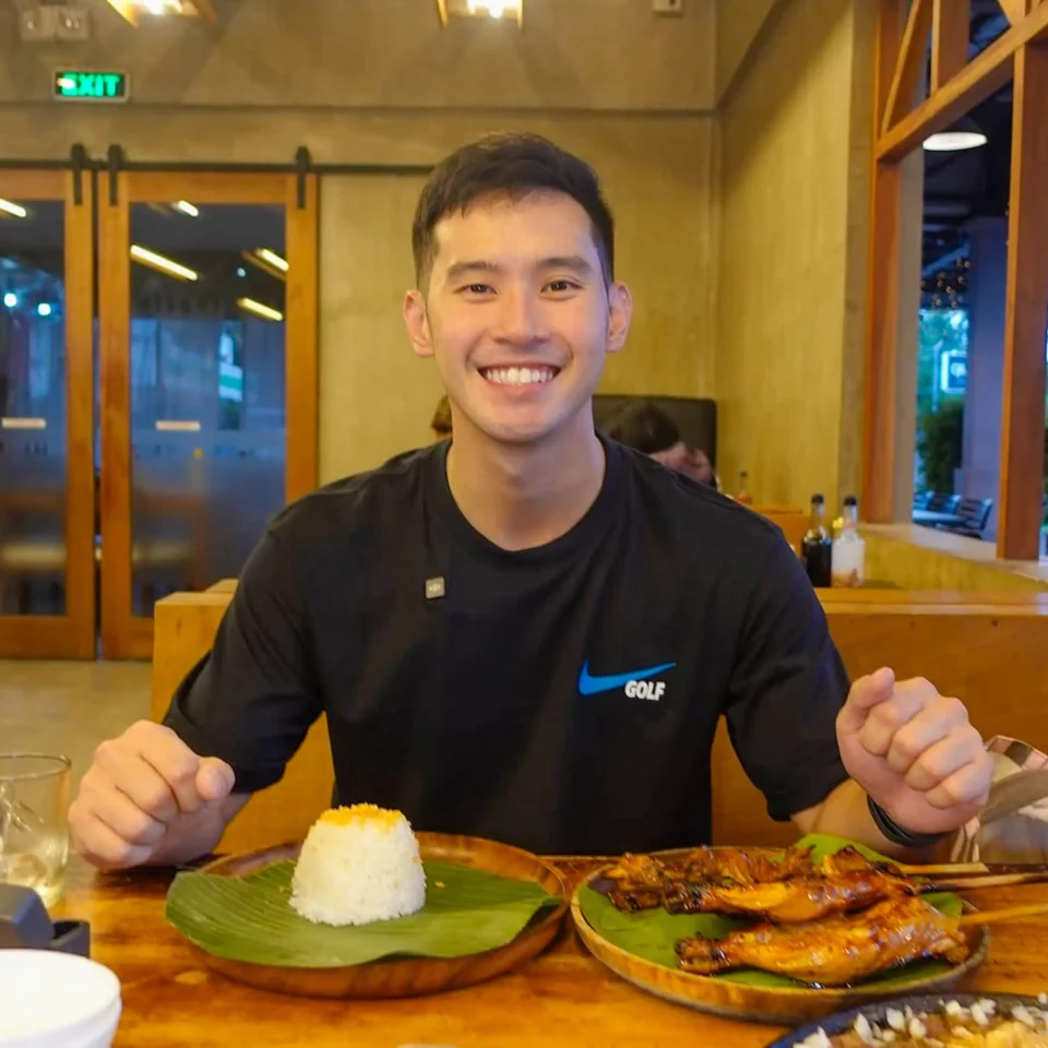 Smiling man in a black shirt sits at a restaurant table with plates of food, including a cone-shaped mound of rice and a platter of grilled skewers in front of him.