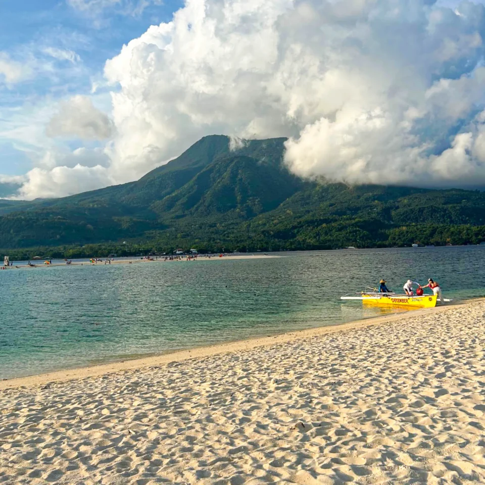 White sand beach in Camiguin Island, Philippines with turquoise water, dramatic volcanic mountains, and large tropical clouds near a peaceful shoreline.