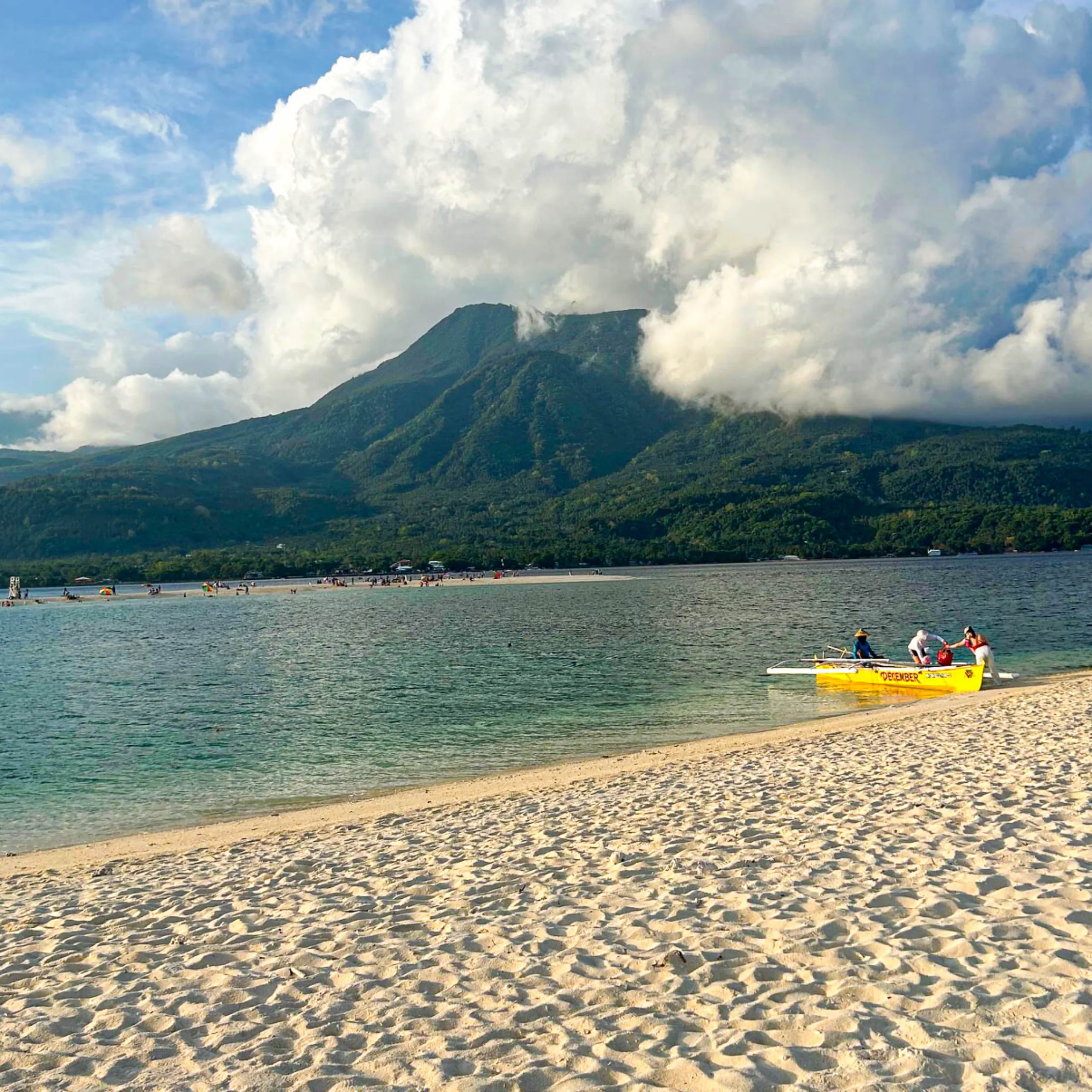 White sand beach in Camiguin Island, Philippines with turquoise water, dramatic volcanic mountains, and large tropical clouds near a peaceful shoreline.
