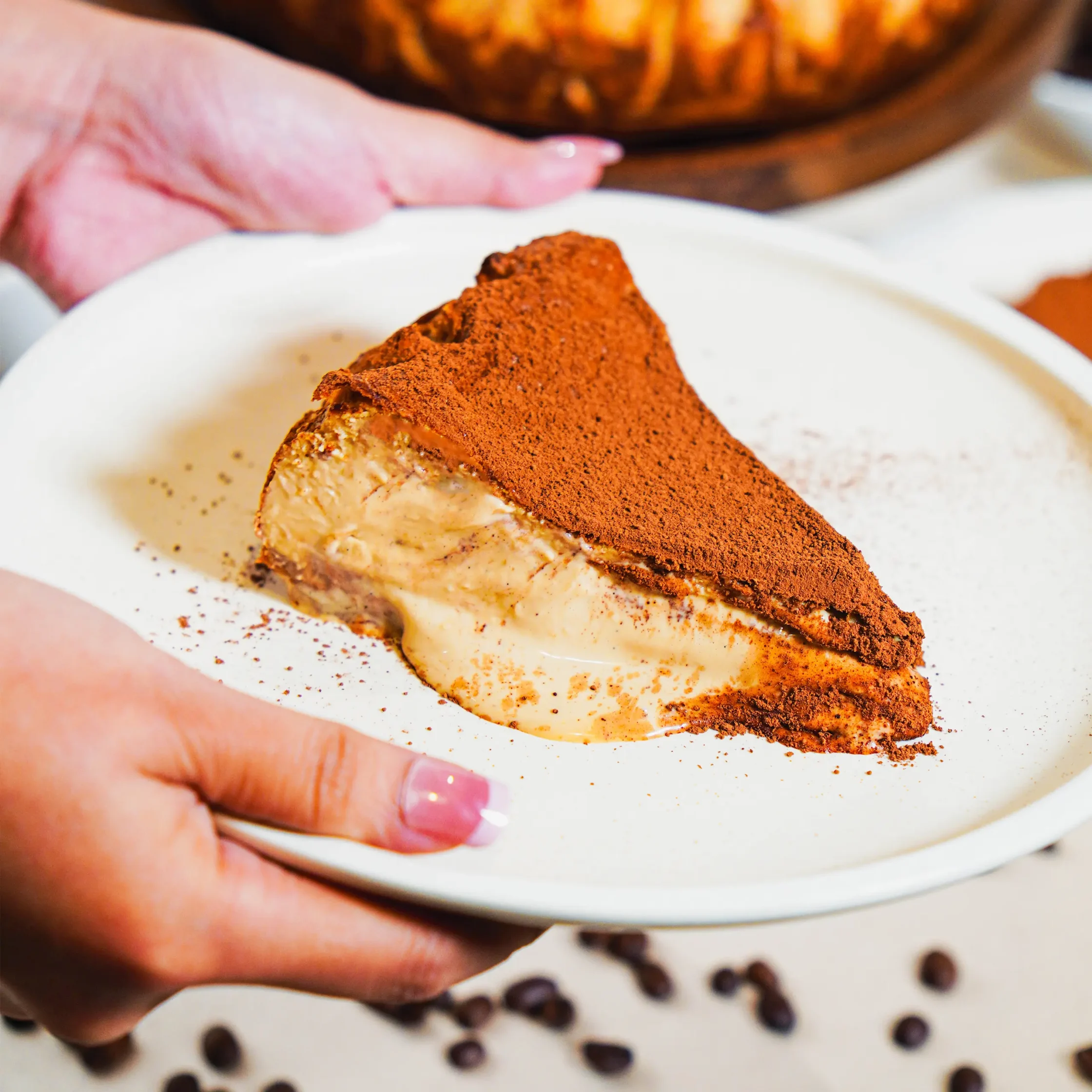 Slice of dessert dusted with cocoa powder on a white plate, a hand resting nearby.