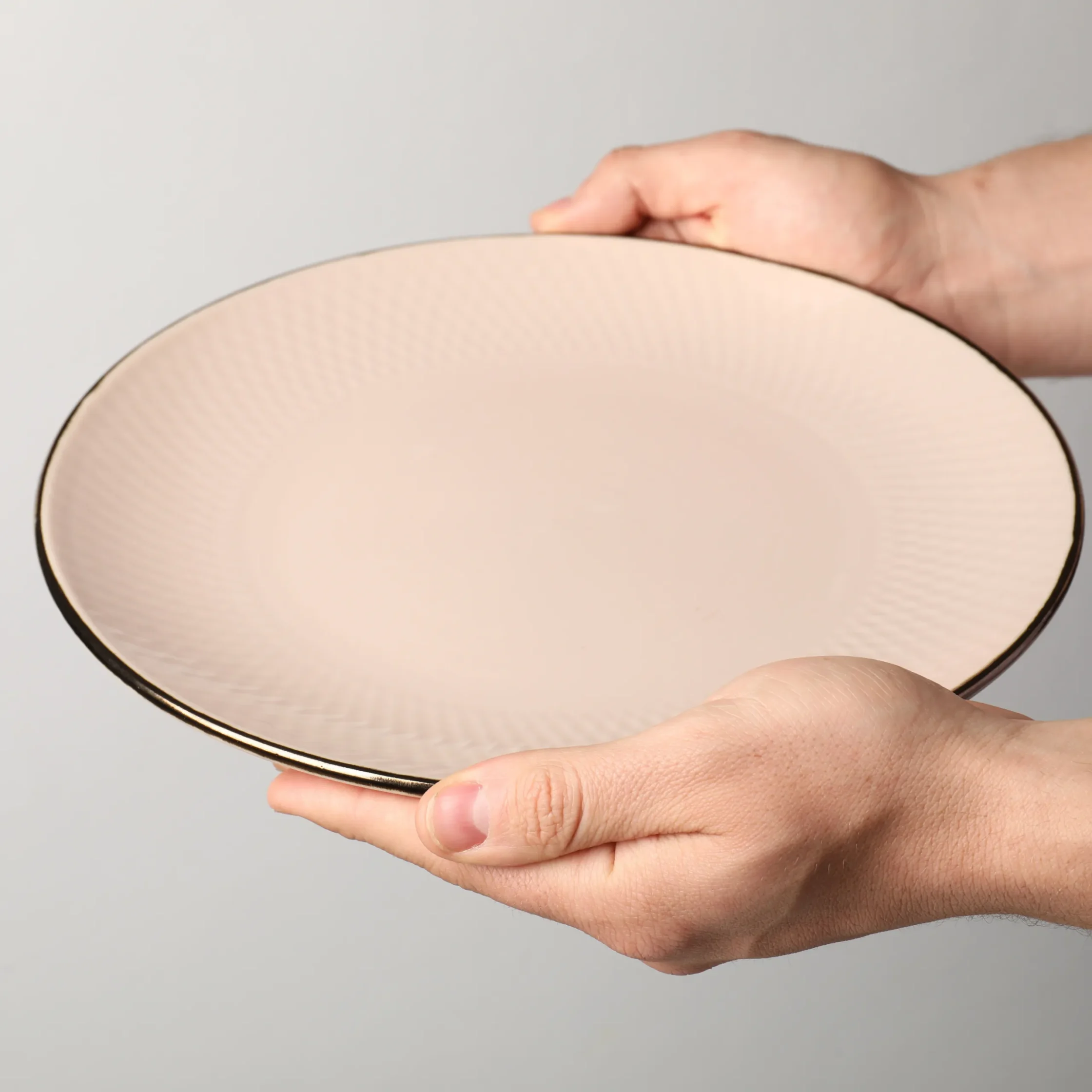 Large ceramic dinner plate with a subtle textured rim, held in two hands against a neutral gray background, commonly used for serving main courses and entrees.