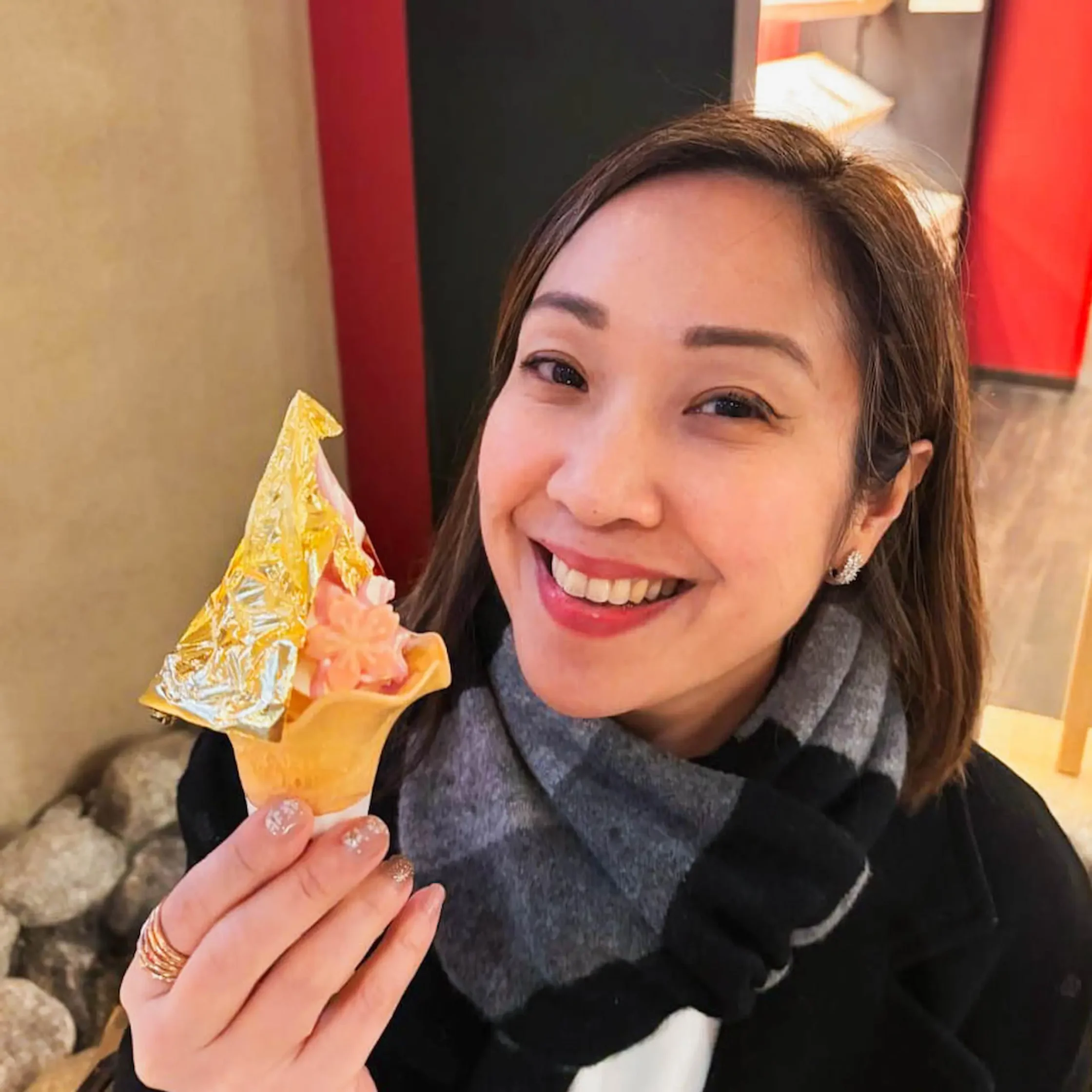 Smiling woman holding a wrapped yellow ice cream cone near her mouth, wearing a dark scarf and gold jewelry.