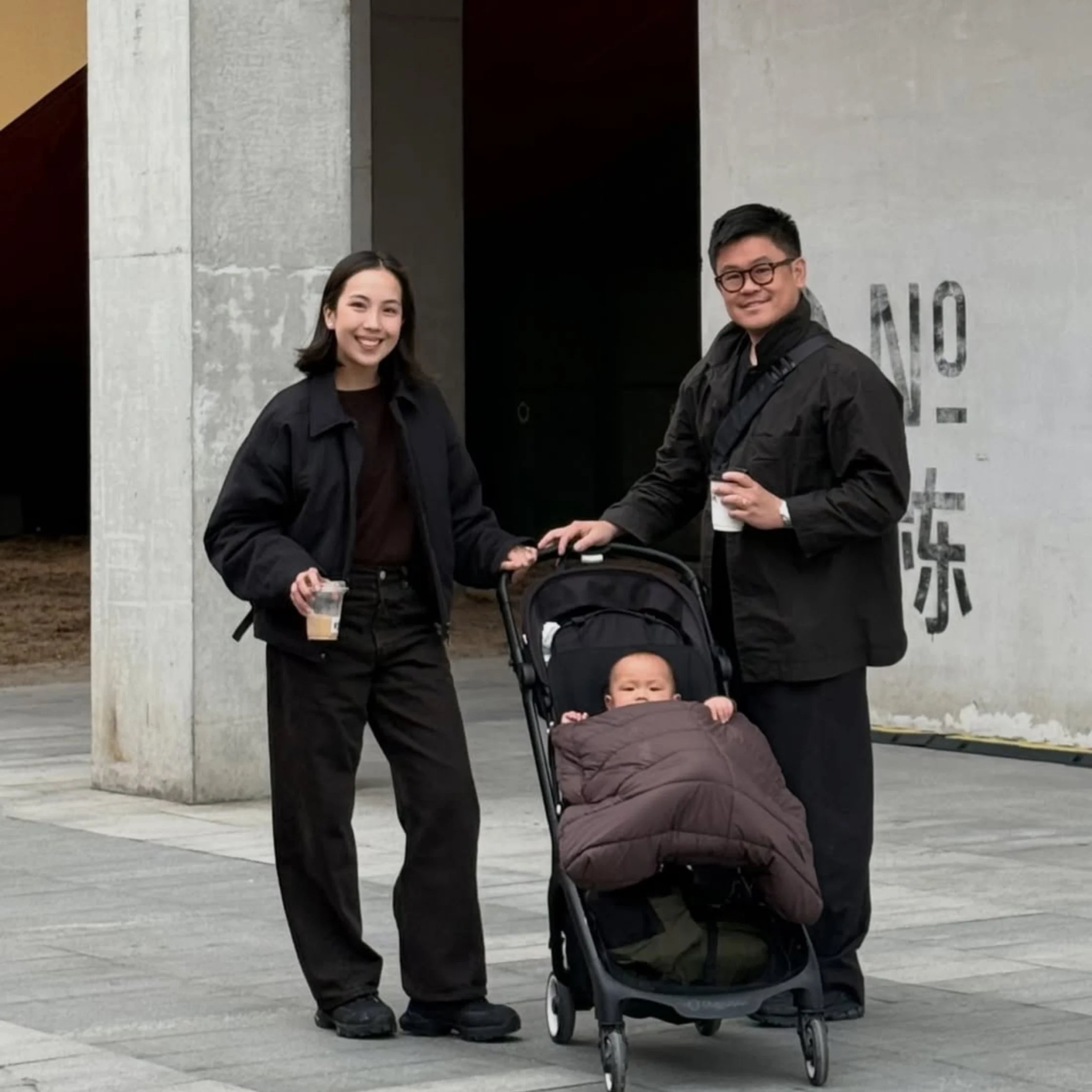 Bryan Kong and Maxine Kong posing with baby Magnus in a stroller outside a modern urban building, stylish family lifestyle portrait