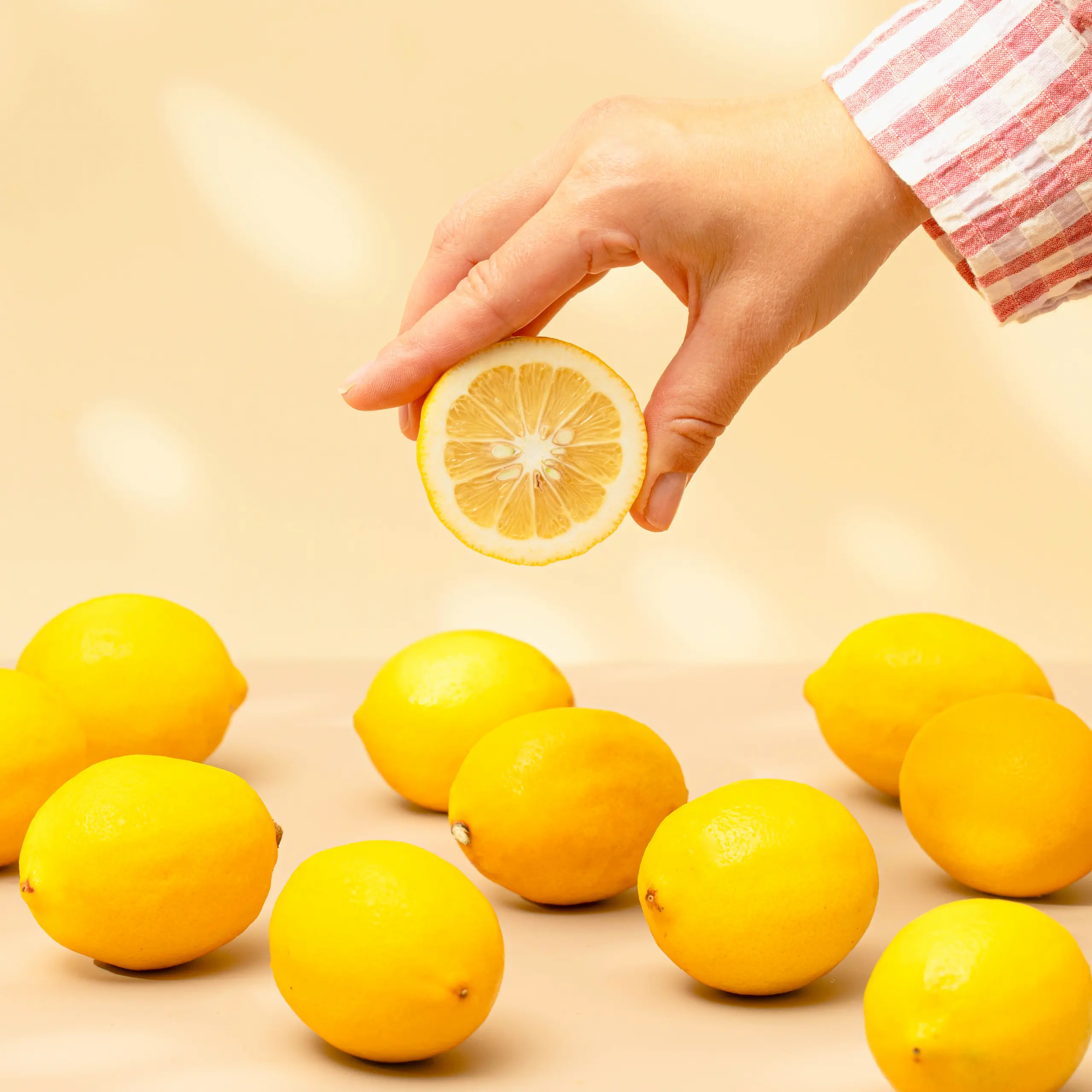 Hand holding a halved lemon over a pale yellow background, with whole lemons lined up below.
