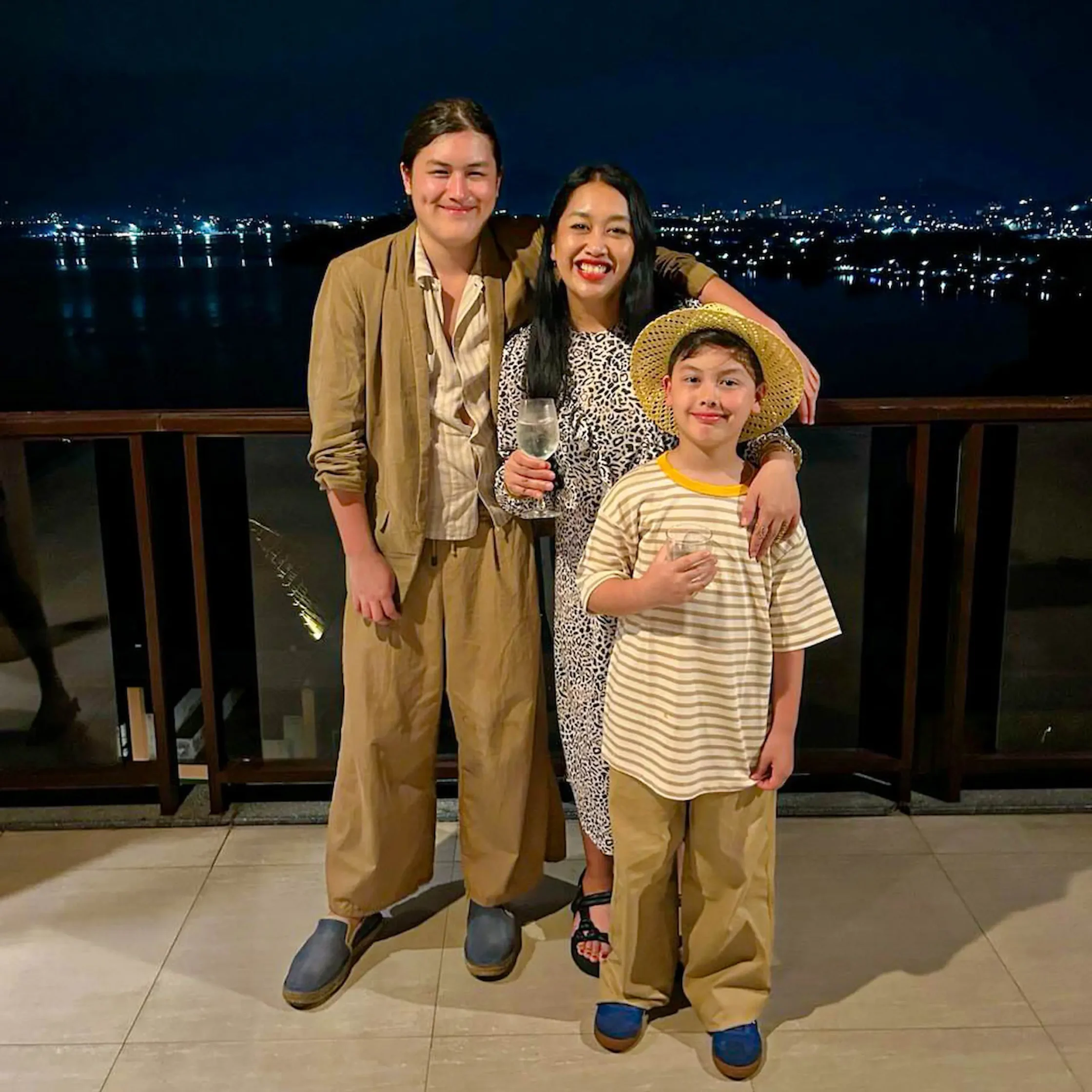 Three people posing on a balcony at night wearing oversized tan pants, smiling at the camera.