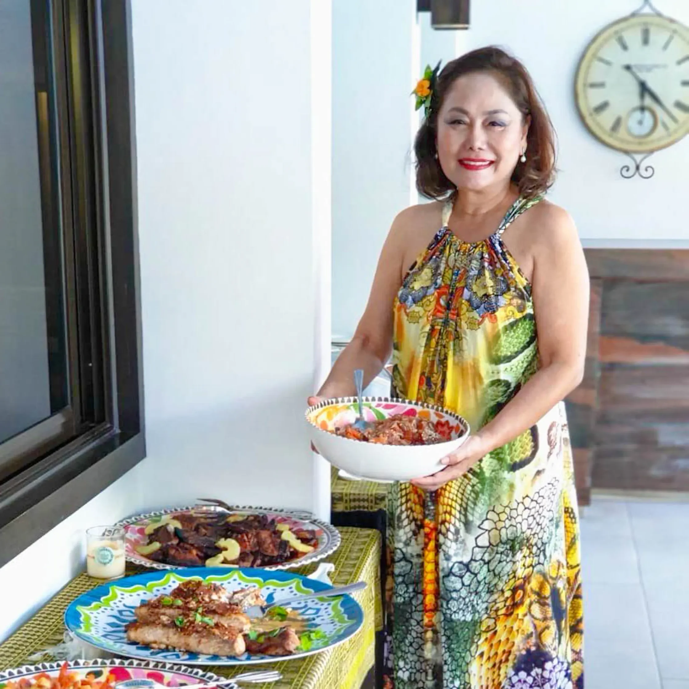 Smiling woman in a colorful dress with a flower in her hair holds a bowl, standing beside a table of assorted food plates.