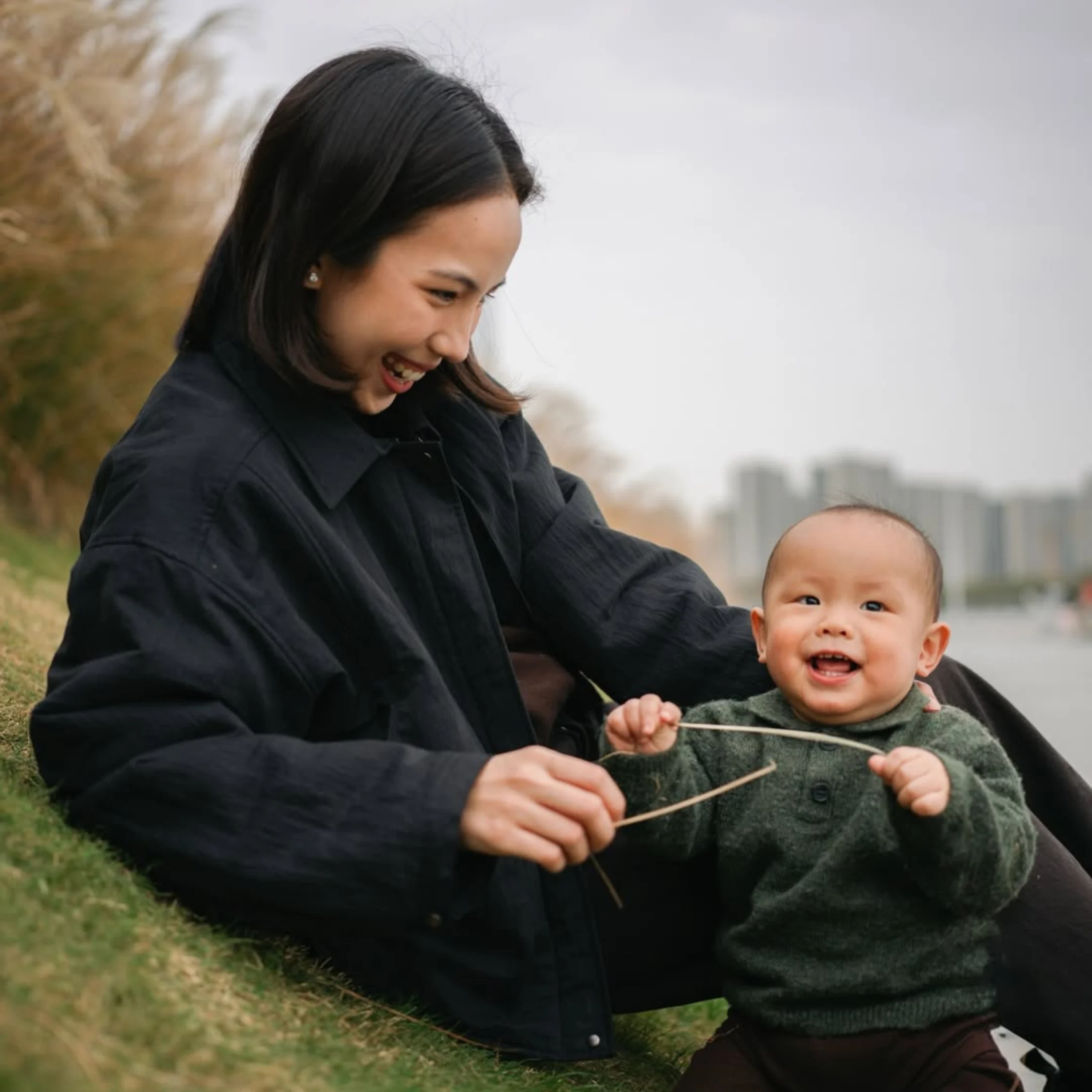 Maxine Kong playing with baby Magnus on a grassy riverside slope, joyful mother and baby outdoor lifestyle portrait