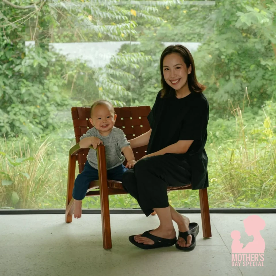 Maxine Kong sitting with baby Magnus indoors beside a large window overlooking lush greenery, candid mother and son portrait