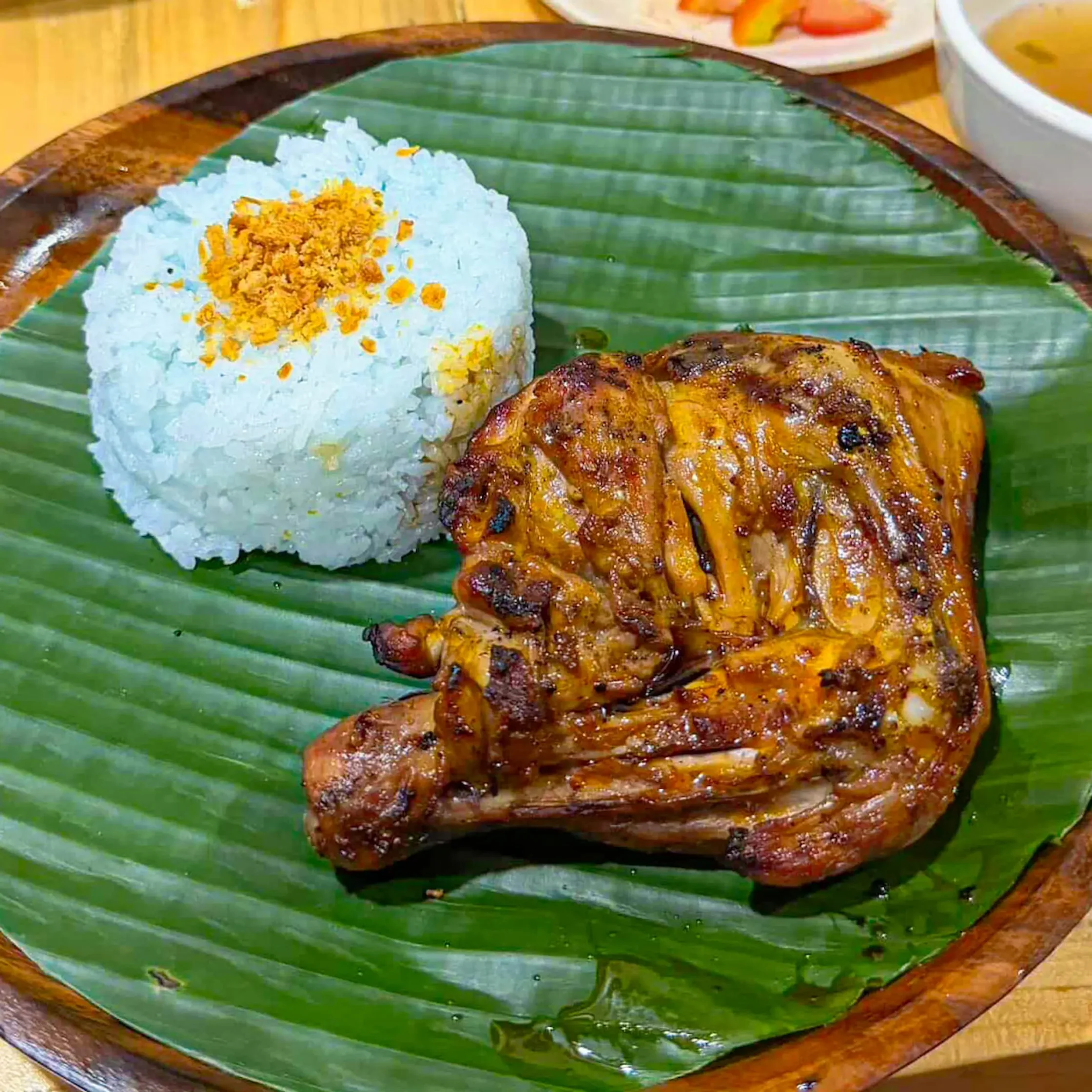 Grilled chicken pieces beside a mound of blue rice topped with crunchy golden bits on a banana-leaf plate.