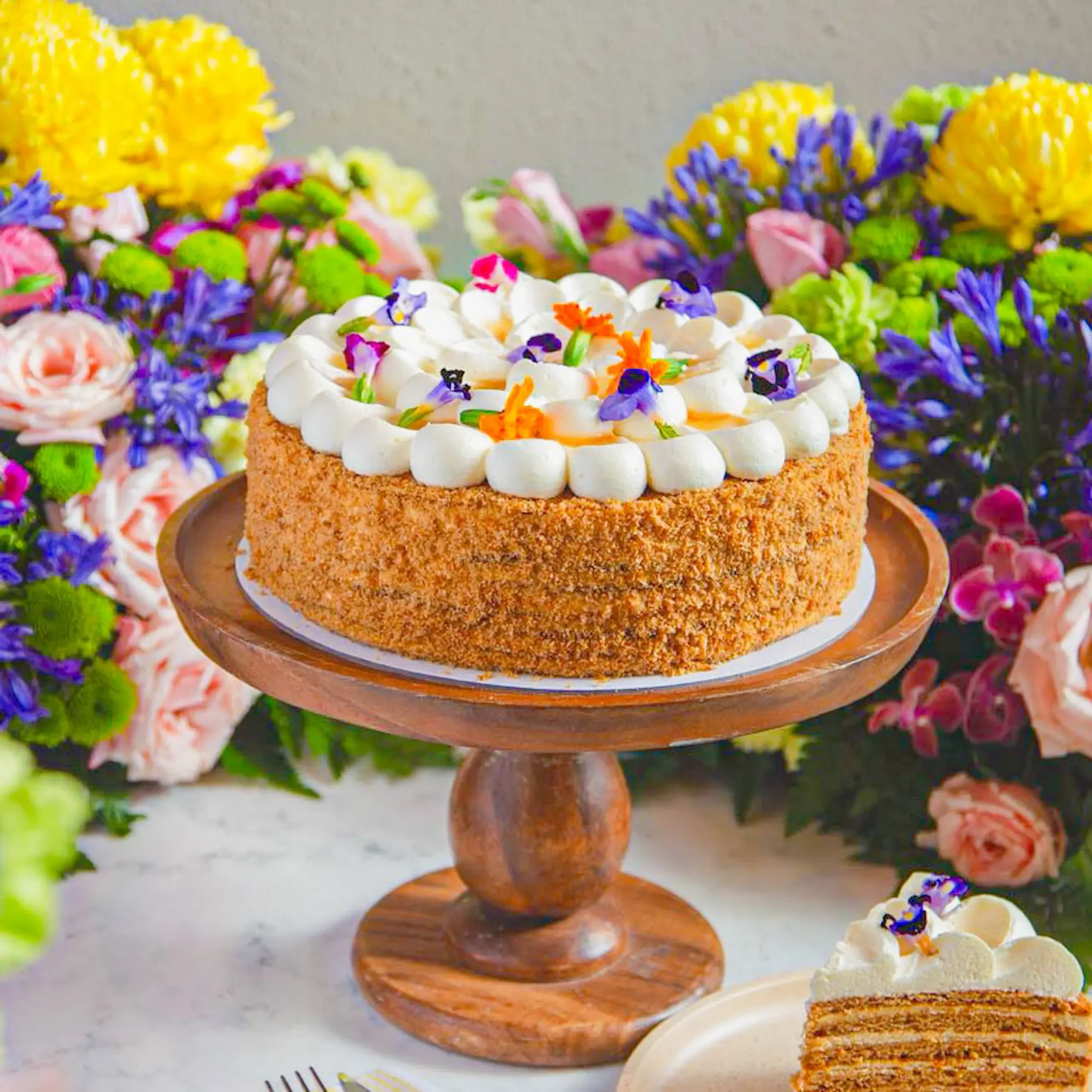 Layered cake with white frosting rosettes and a crunchy crust on a wooden stand, with a vivid flower backdrop.