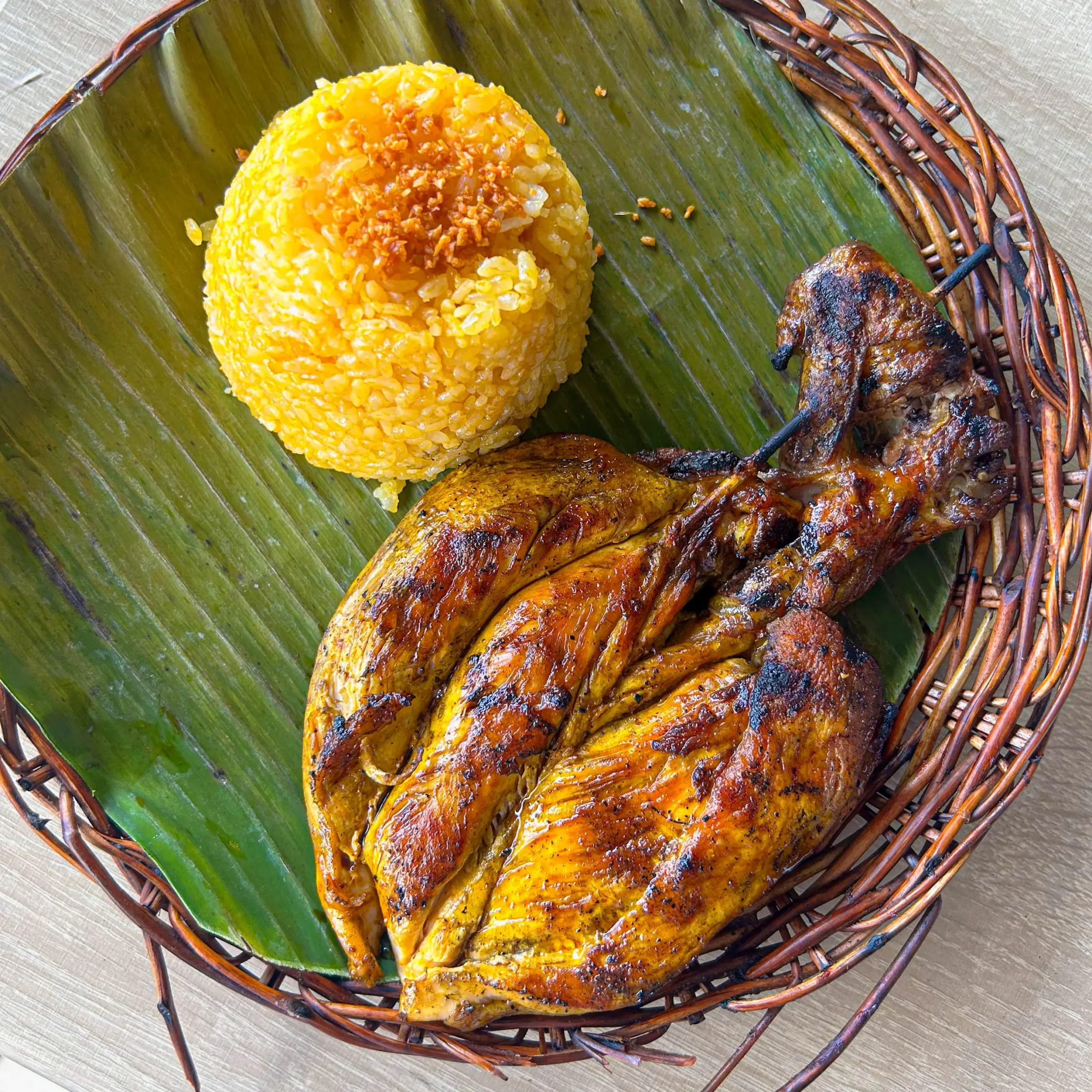 Yellow fried rice served on a banana leaf in a woven basket with grilled sausages beside it.