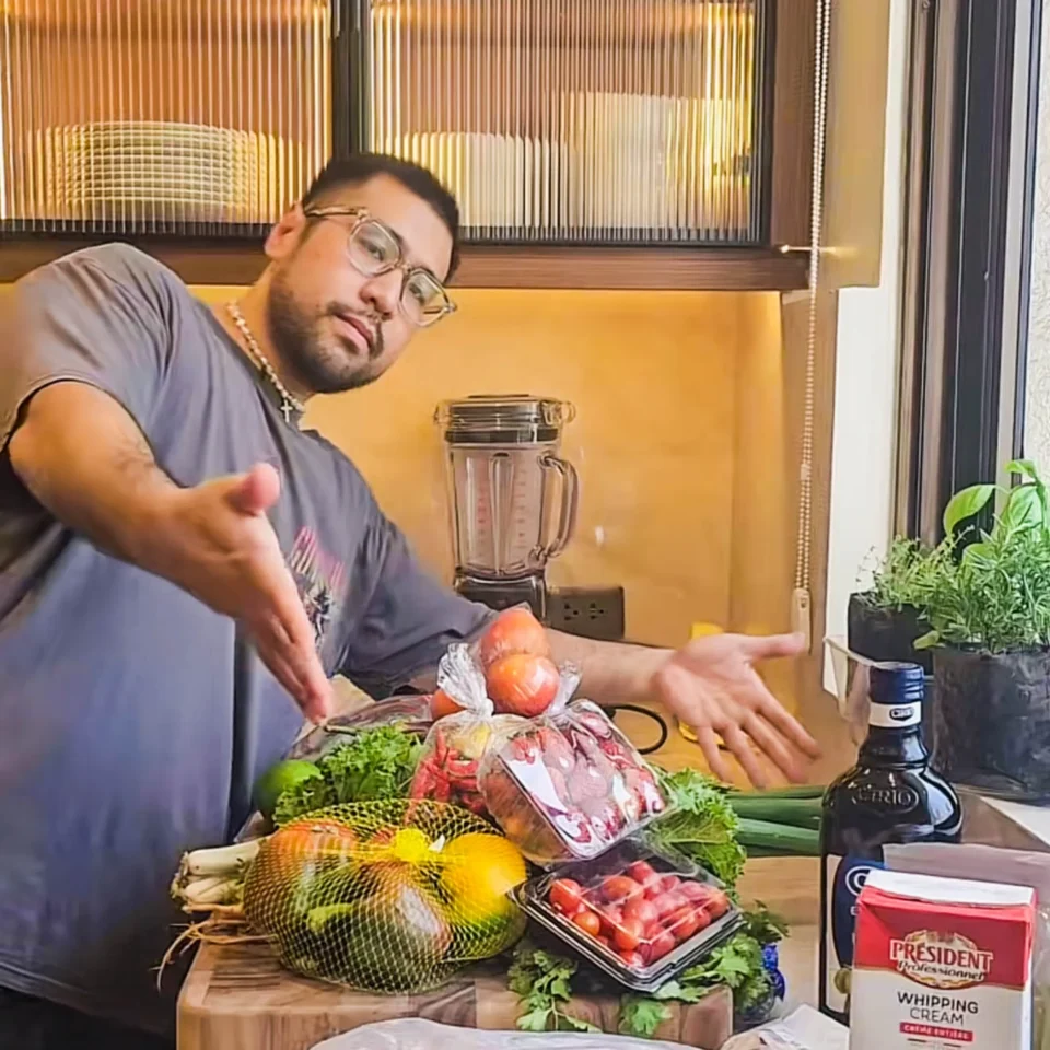 A man wearing glasses gestures toward an assortment of fresh produce, herbs, and cooking ingredients displayed on a kitchen counter beside a window. private chef geron altamirano