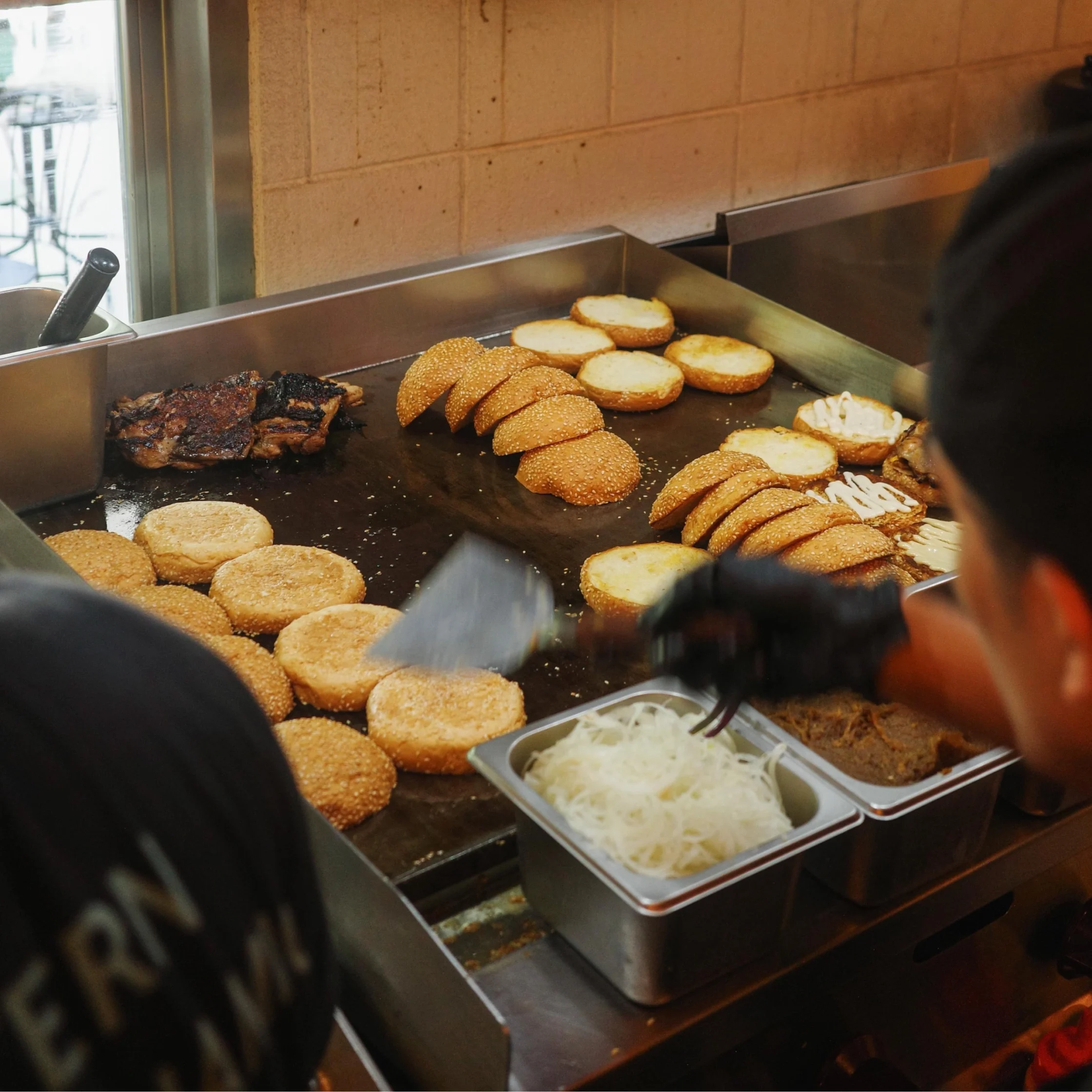 Hamburger buns and beef patties cooking on a griddle in a restaurant kitchen.